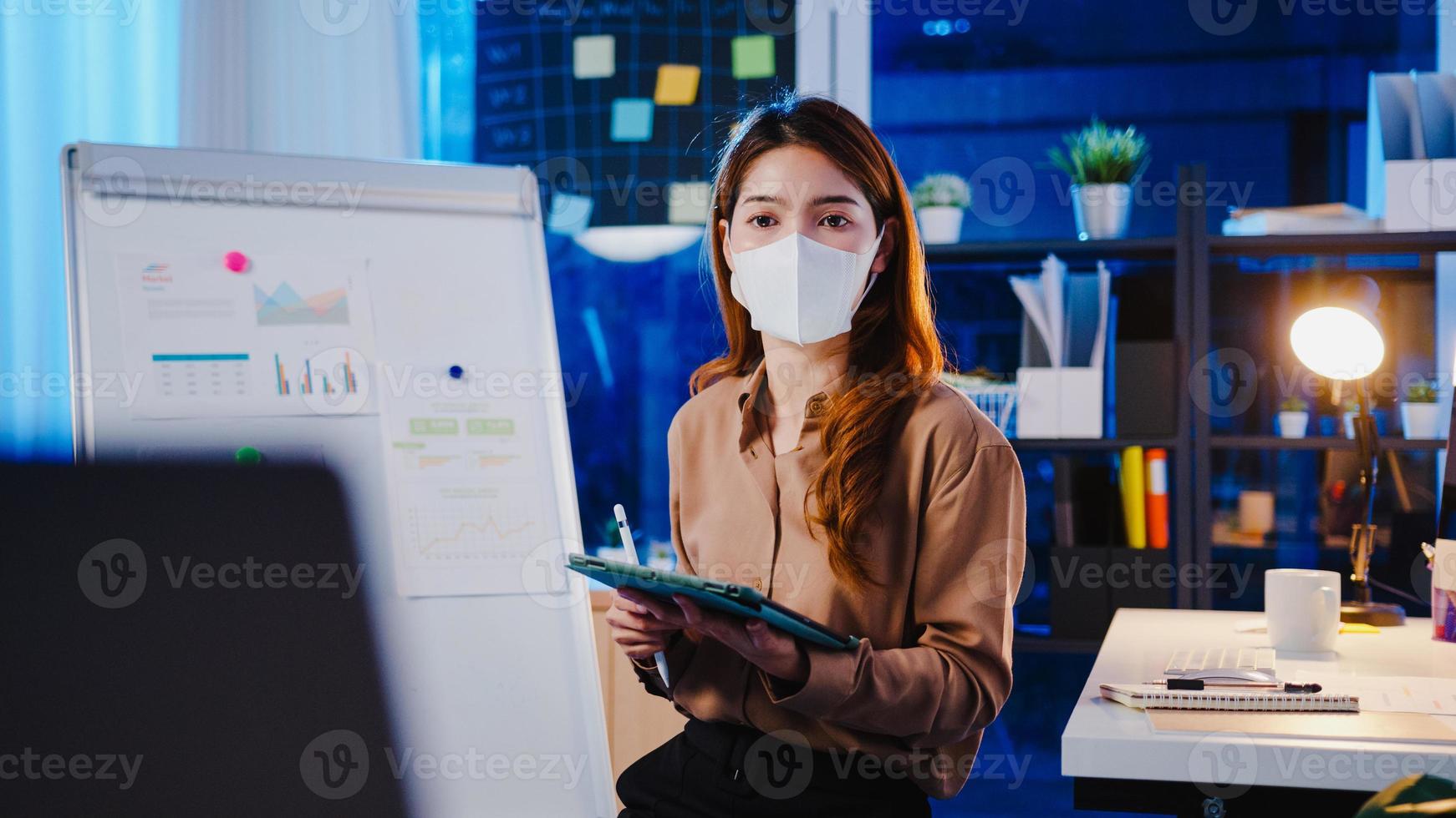 asiatisk affärskvinna bär ansiktsmask för social distansering i ny normal för virusförebyggande presentation till kollega om plan i videosamtal medan du arbetar på kontorsnatt. livsstil efter coronaviruset. foto