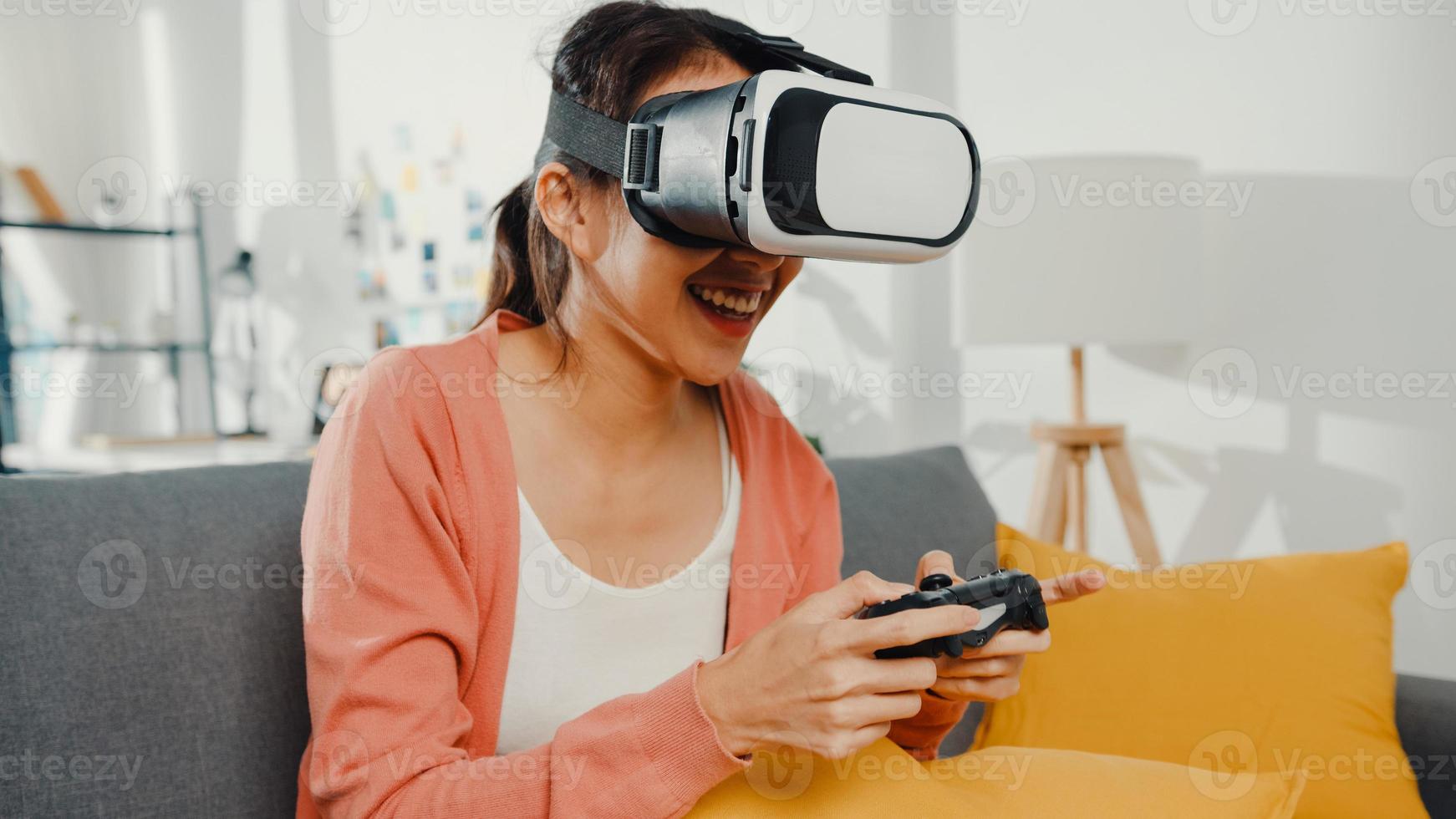 asiatisk dam bär headsetglasögon av virtuell verklighet spela joystick -spel på soffan i vardagsrummet hemma. stanna hemma covid karantän, tänk om verkligheten, vr hemma, vr teknik för framtida koncept. foto