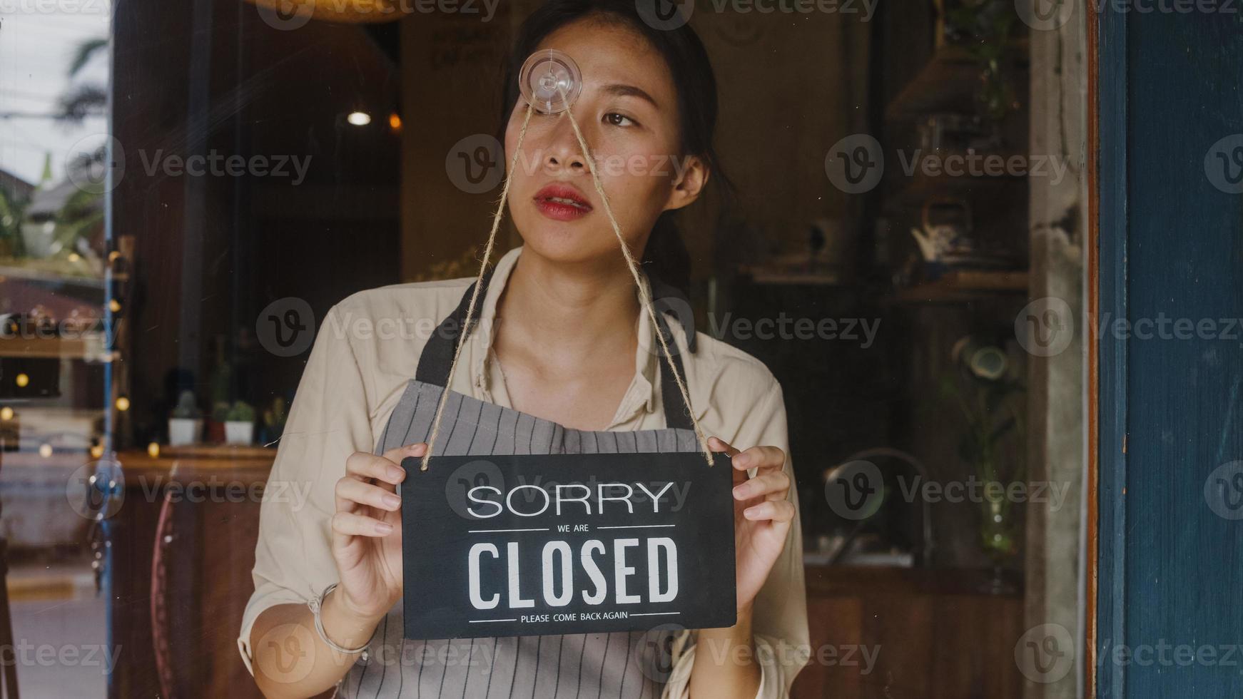 ung asiatechef tjej vänder en skylt från öppen till stängd skylt på glasdörrkafé efter koronantillfällning av coronavirus. ägare småföretag, mat och dryck, affärsfinansiell kriskoncept. foto