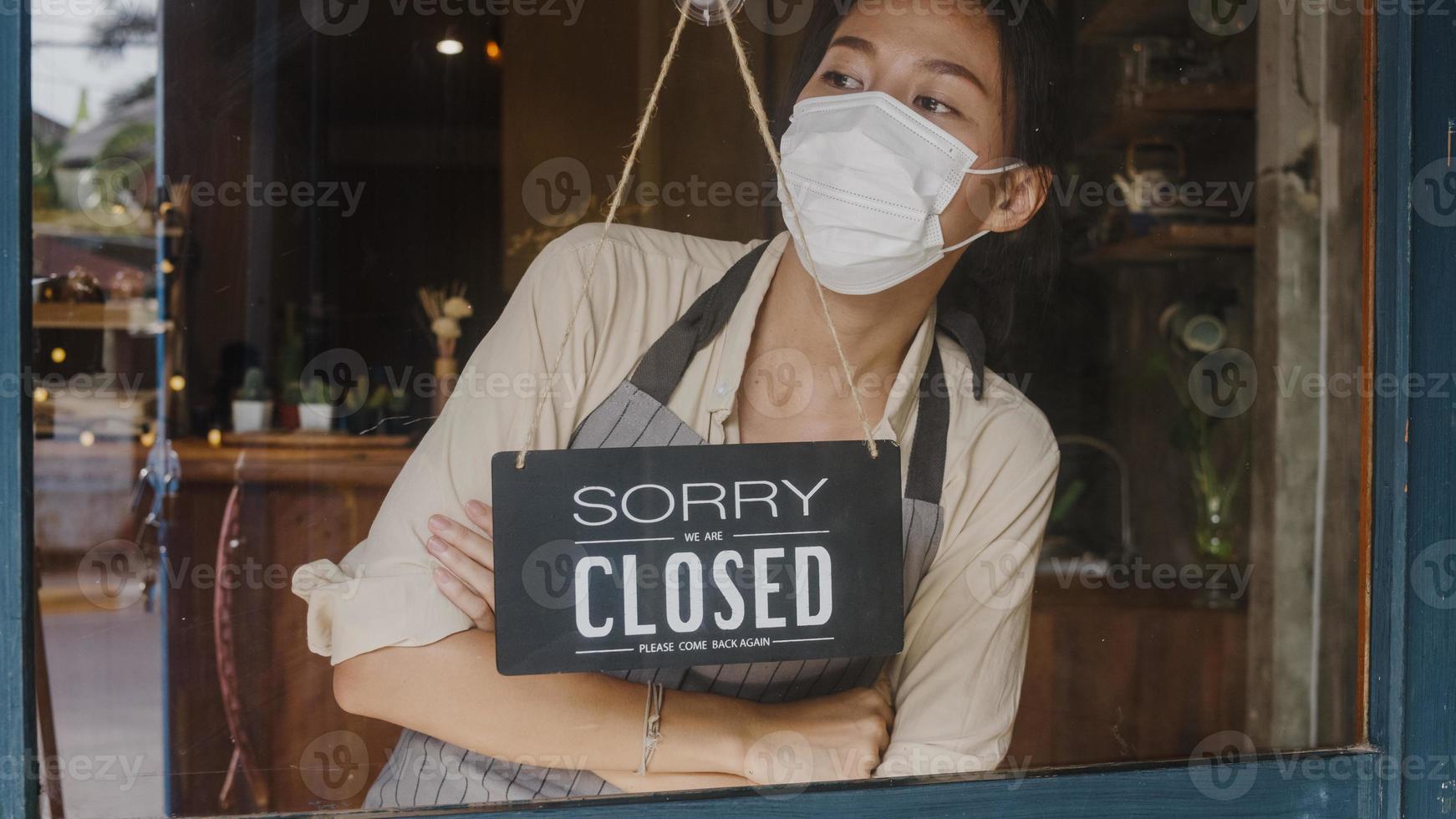 ung asiatisk tjej bär ansiktsmask som vänder en skylt från öppen till stängd skylt på glasdörrkafé efter koronaklockan. ägare småföretag, mat och dryck, affärsfinansiell kriskoncept foto