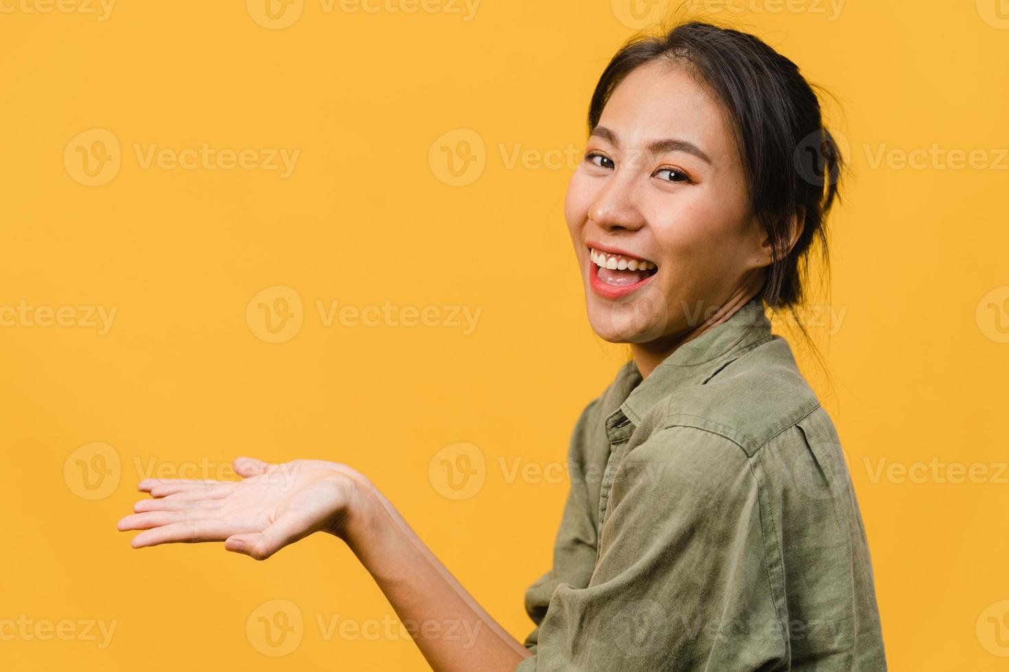 porträtt av ung asiatisk dam som ler med glatt uttryck, visar något fantastiskt på tomt utrymme i vardagsduk och tittar på kameran isolerad över gul bakgrund. ansiktsuttryck koncept. foto