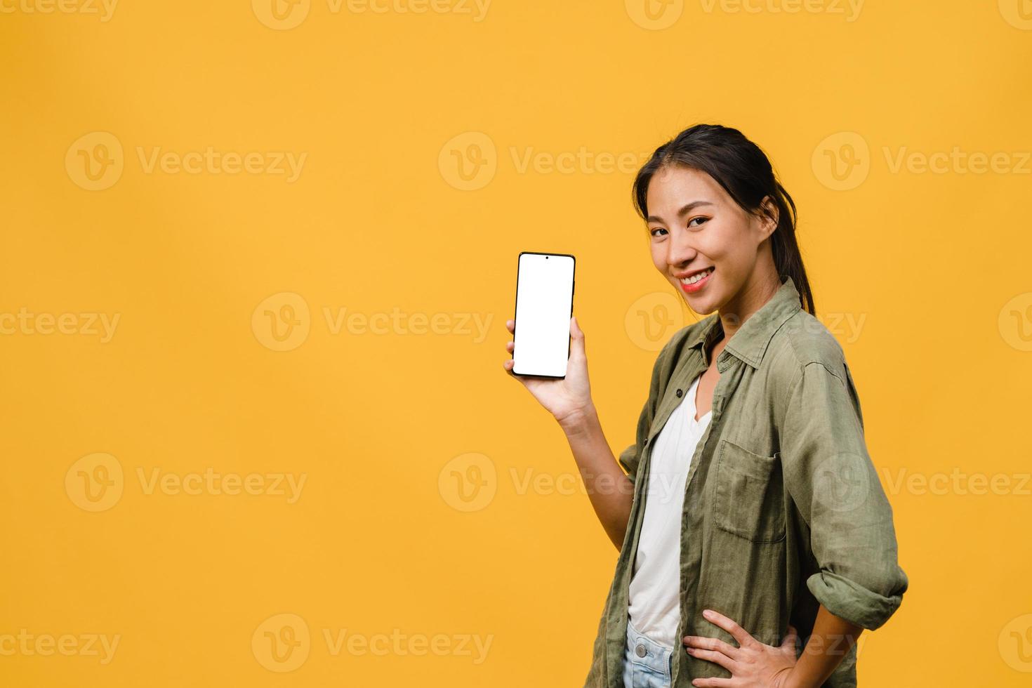 ung asiatisk dam visar tom smartphone -skärm med positivt uttryck, ler brett, klädd i vardagskläder som känner lycka på gul bakgrund. mobiltelefon med vit skärm i kvinnlig hand. foto
