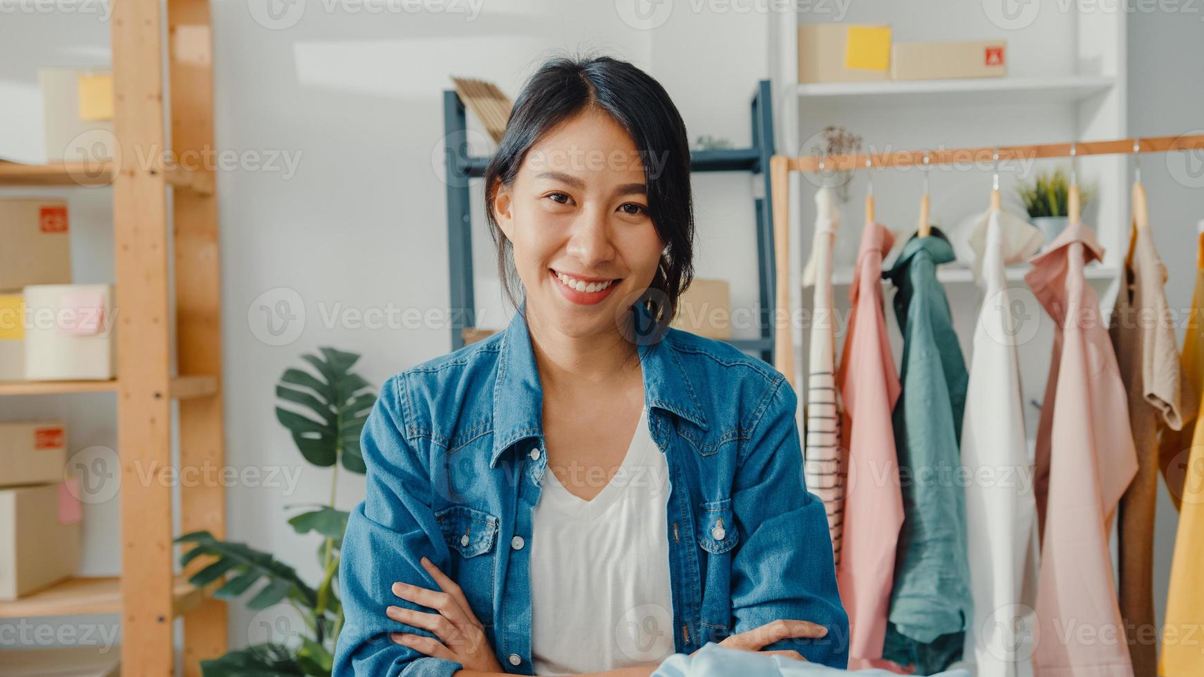 porträtt av ung asiatisk dammodedesigner som känner ett lyckligt leende, korsade armar och ser till kameran medan du arbetar med klädbutik på hemmakontoret. småföretagare, online marknadsföringskoncept. foto