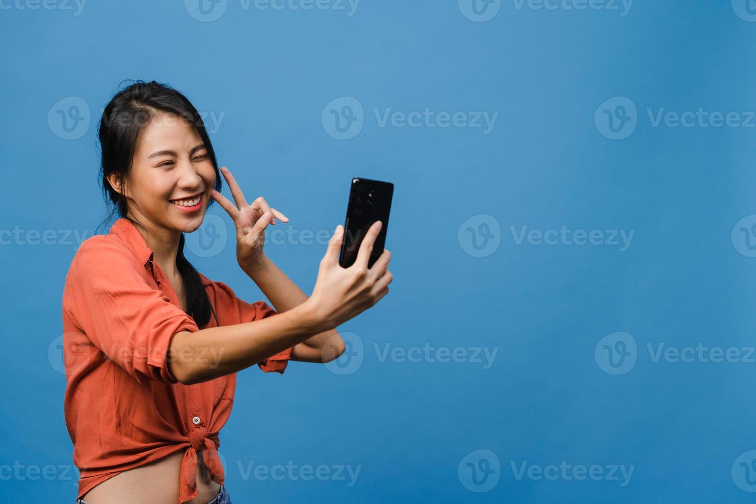 leende bedårande asiatisk kvinna som gör selfie -foto på smart telefon med positivt uttryck i vardagskläder och stativ isolerad på blå bakgrund. glad förtjusande glad kvinna jublar över framgång. foto