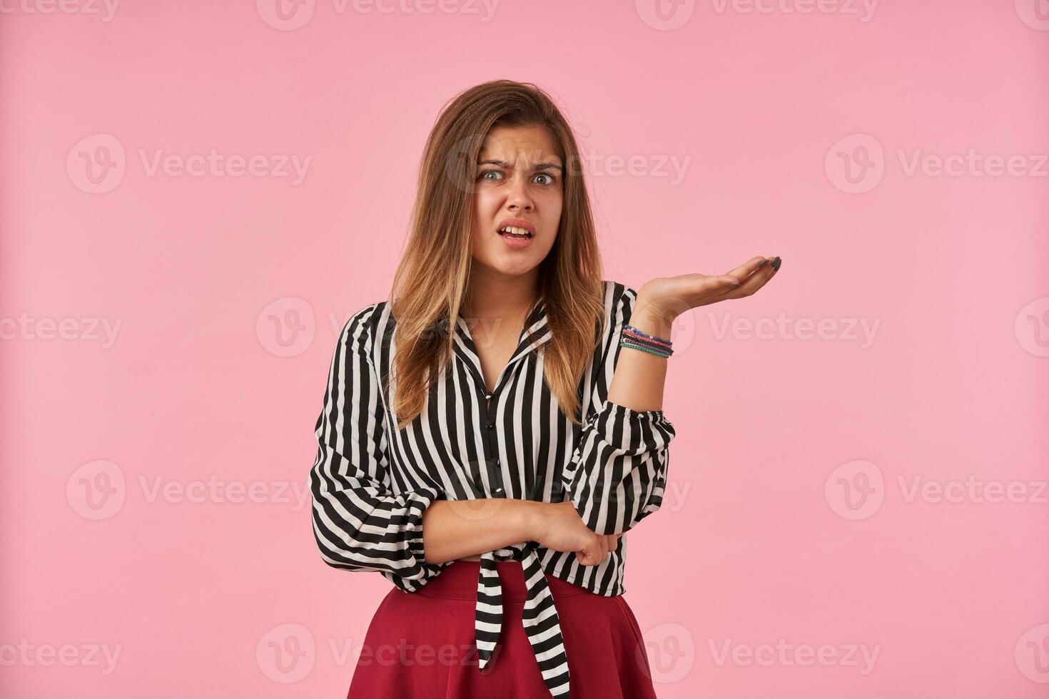 generad ung brun håriga grönögd kvinna grimaserande henne ansikte och ser missnöjt på kamera, stående mot rosa bakgrund med Uppfostrad handflatan foto