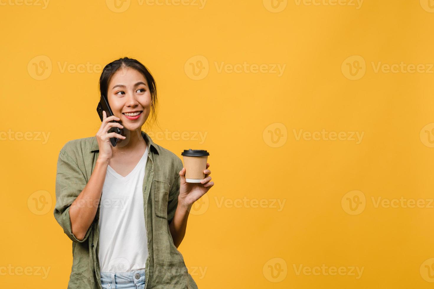 ung asiatisk dam pratar i telefon och håller kaffekoppen med positivt uttryck, ler brett, klädd i avslappnad trasa som känner lycka och står isolerad på gul bakgrund. ansiktsuttryck koncept. foto