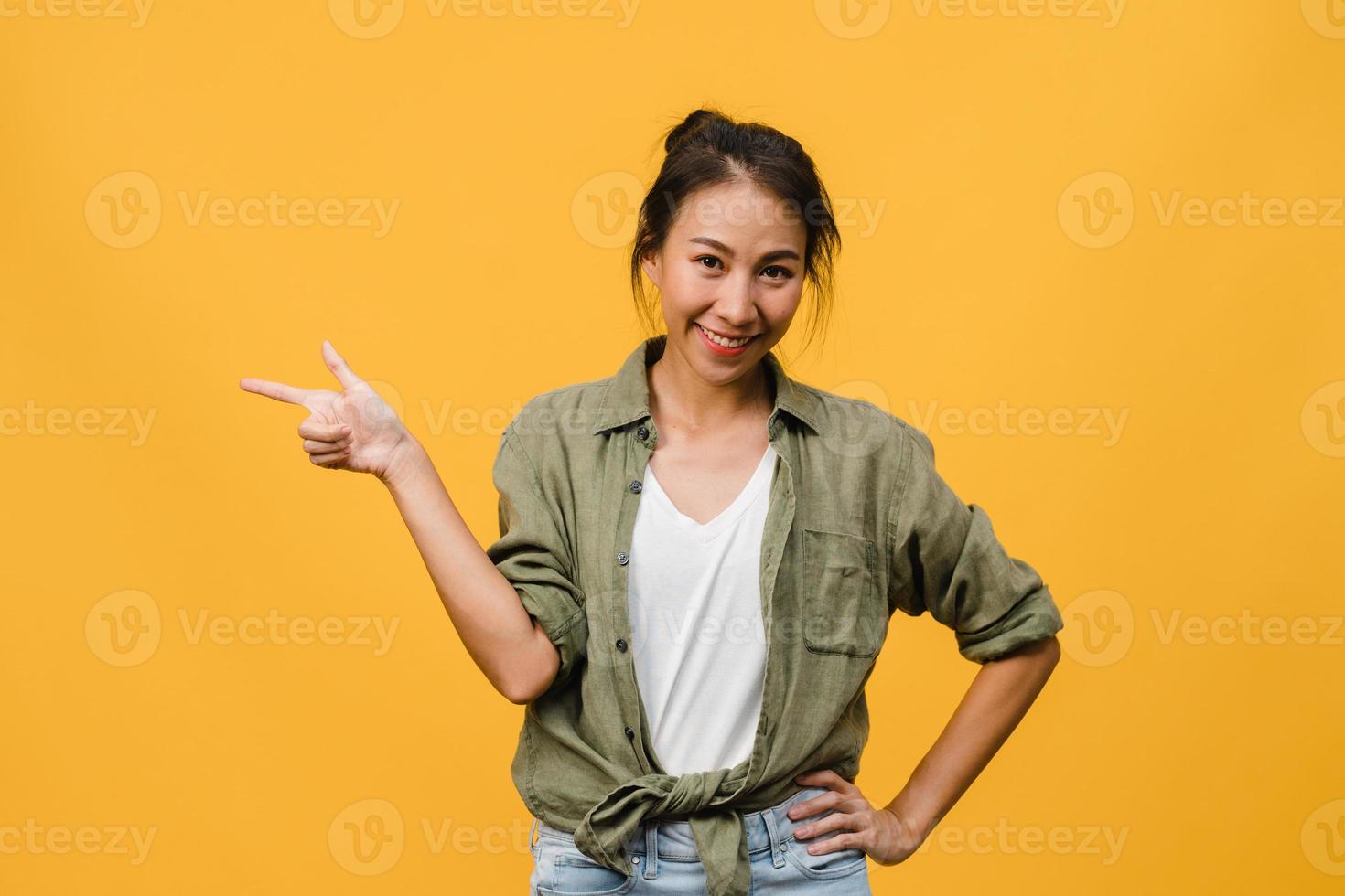 porträtt av ung asiatisk dam som ler med glatt uttryck, visar något fantastiskt på tomt utrymme i vardagsduk och tittar på kameran isolerad över gul bakgrund. ansiktsuttryck koncept. foto
