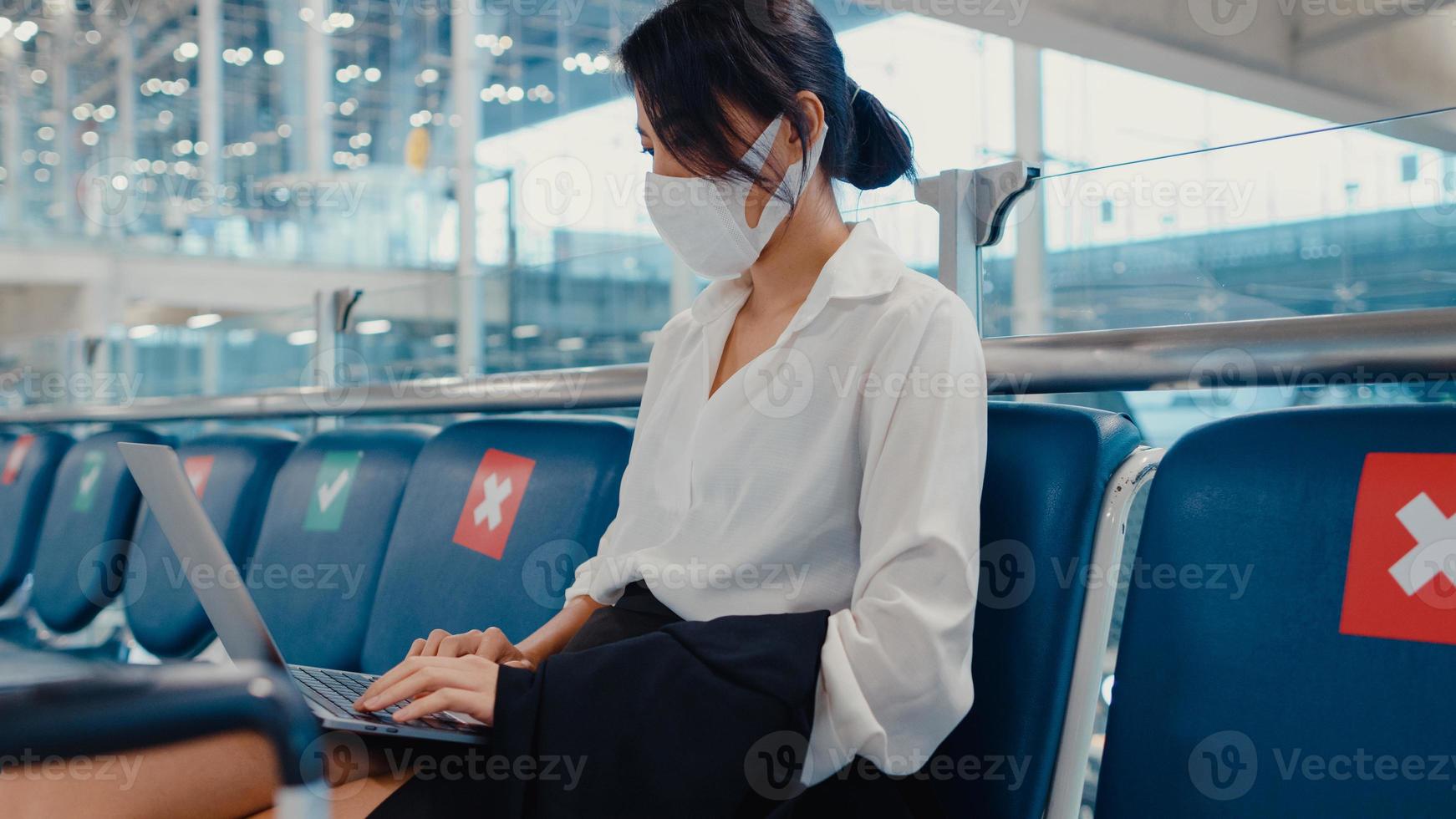 asiatisk affärsresande damresenär bär ansiktsmask som sitter i bänken använder bärbar dator för arbete mellan väntan på flygning i terminalen på flygplatsen. affärsresependlare i covid -pandemi, affärsresekoncept. foto