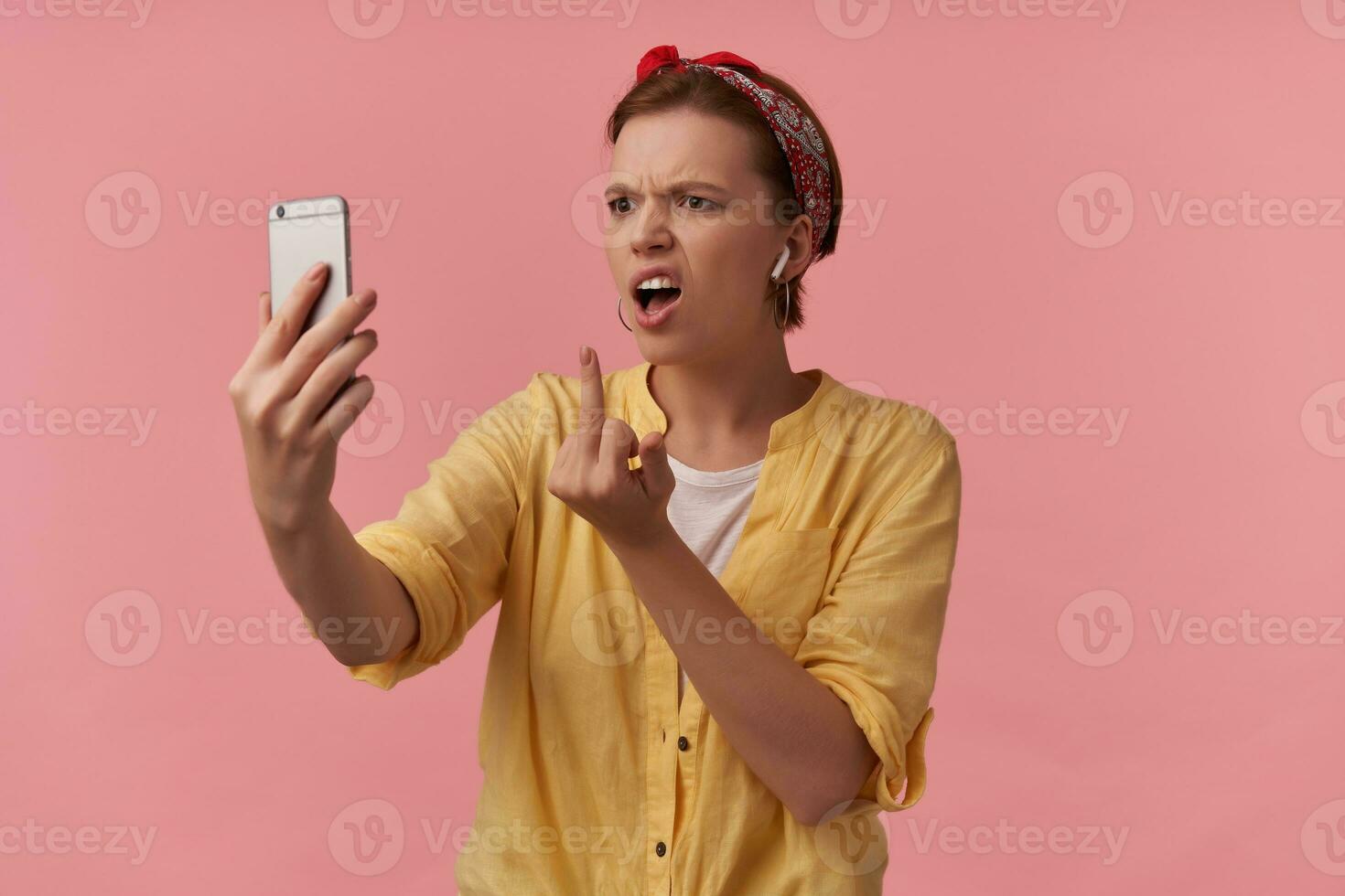 bild europeisk brun håriga ungdom kvinna vit t-shirt och gul skjorta och röd bandana Framställ på själv skott känsla oförskämd galen arg olycklig mitten finger ser på telefon isolerat över rosa bakgrund foto