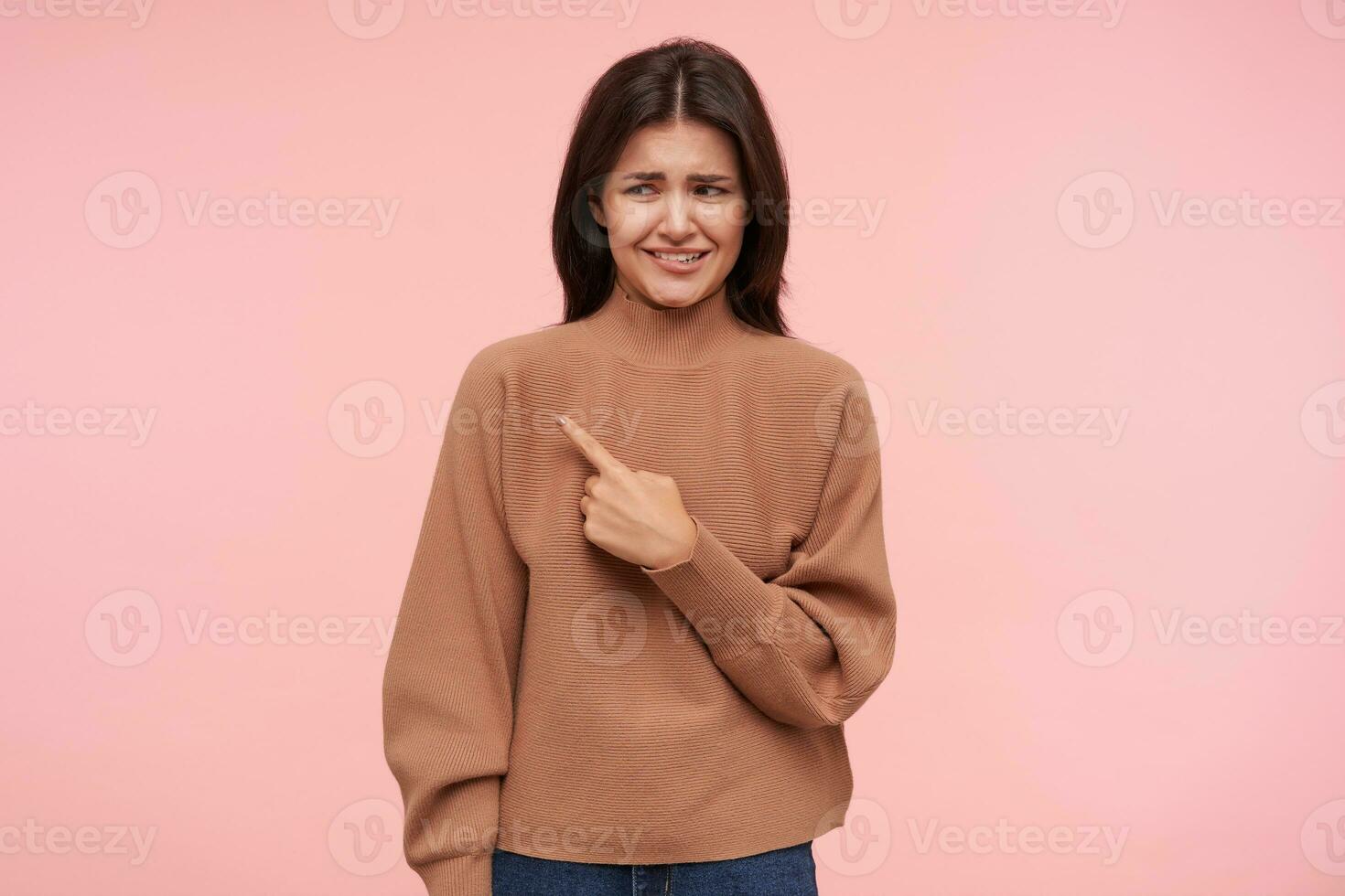 studio skott av förvirrad ung brunett lady grimaserande henne ansikte medan pekande åt sidan med Uppfostrad index finger, stående över rosa bakgrund i brun polotröja foto