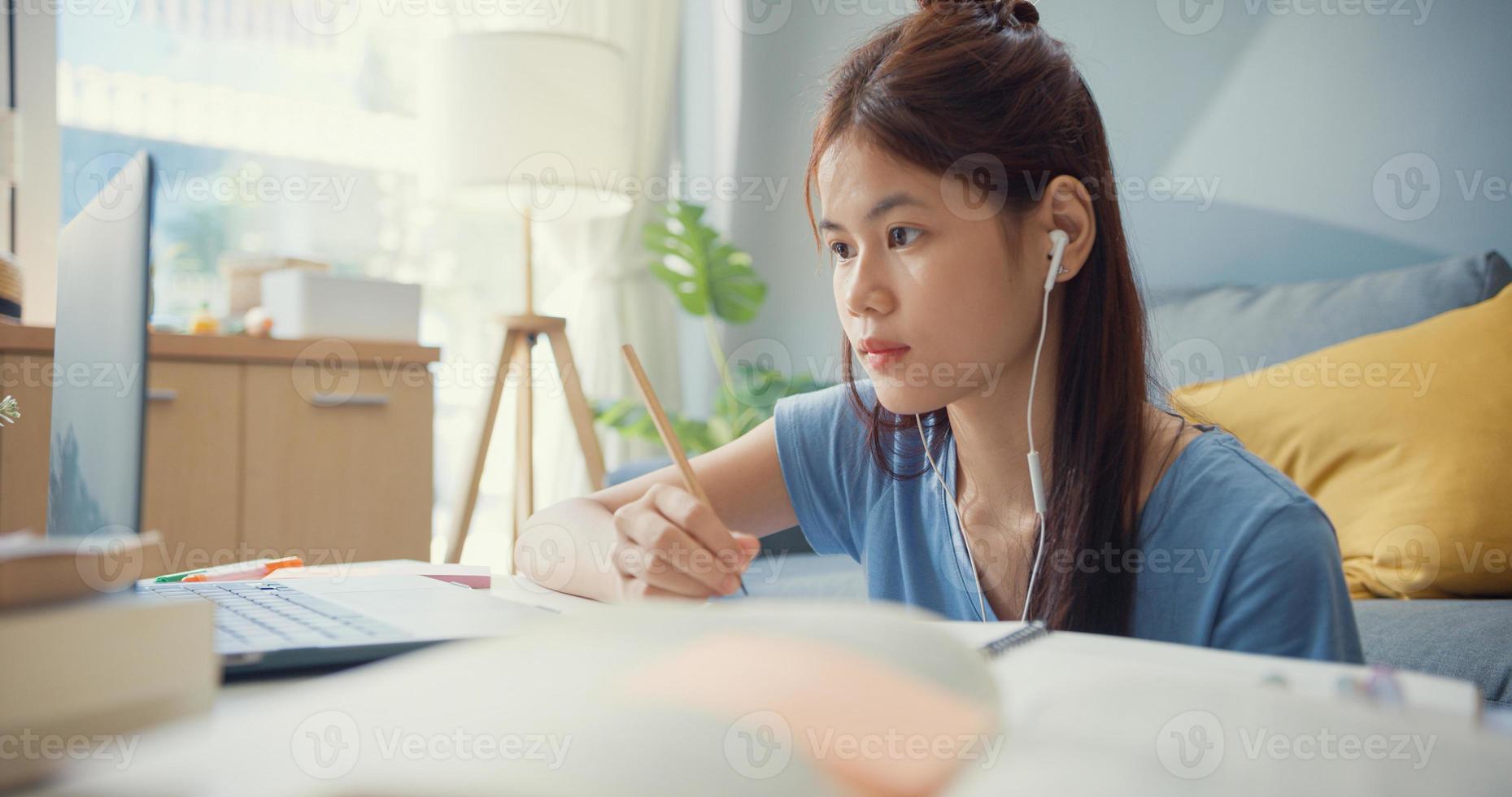 ung asiatisk tjej tonåring med slitage hörlurar använda bärbar dator lära sig online skriva föreläsning anteckningsbok i vardagsrummet hemma. isolera utbildning online e-learning coronavirus-pandemikoncept. foto