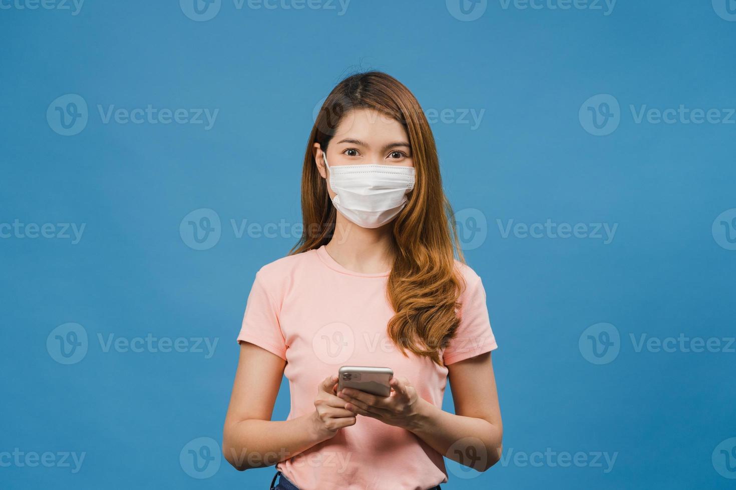 ung asiatisk tjej som bär medicinsk ansiktsmask med mobiltelefon med klädda i vardagskläder isolerad på blå bakgrund. självisolering, social distansering, karantän för förebyggande av corona-virus. foto