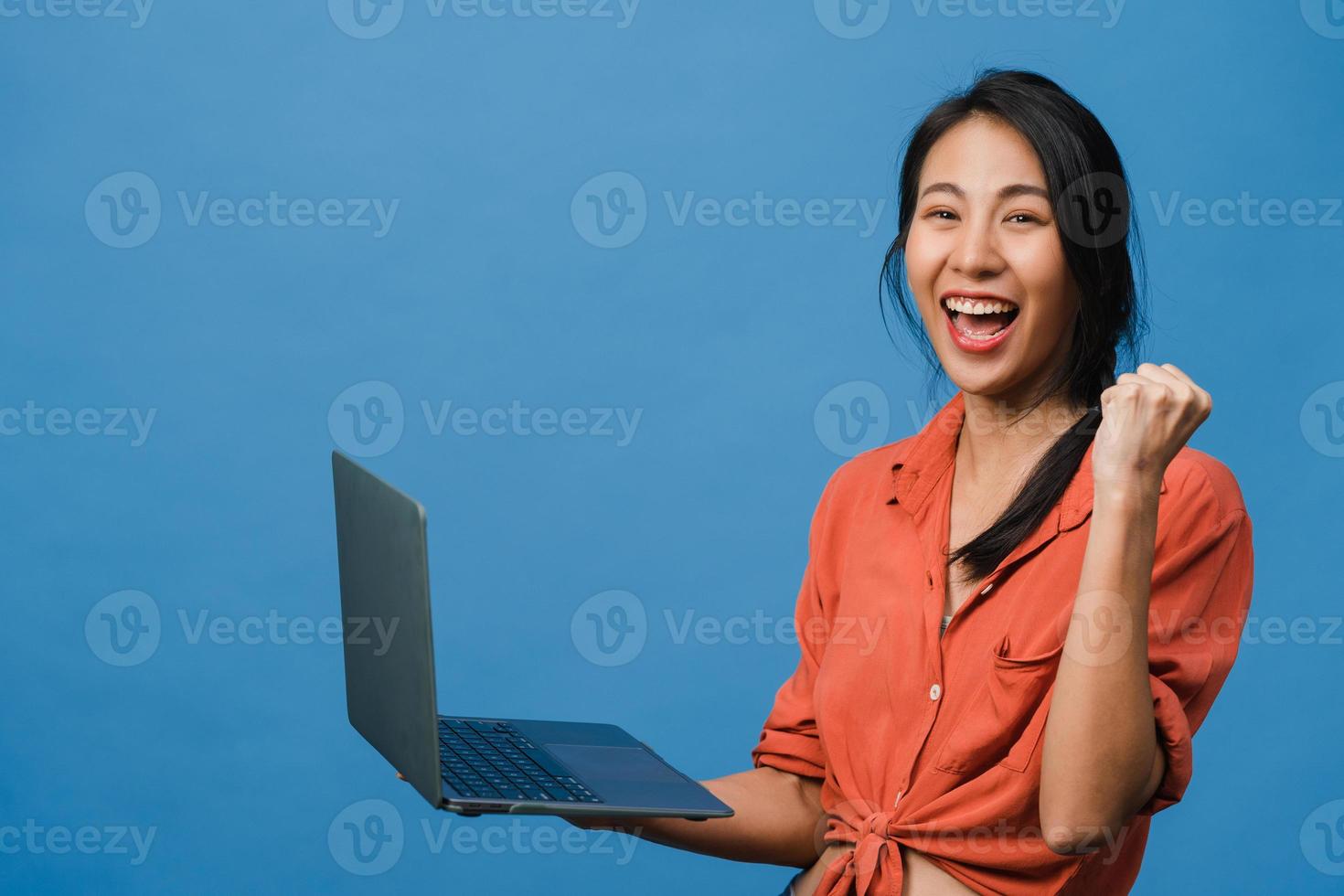 ung asiatisk dam som använder bärbar dator med positivt uttryck, ler brett, klädd i vardagskläder som känner lycka och står isolerad på blå bakgrund. glad förtjusande glad kvinna jublar över framgång. foto