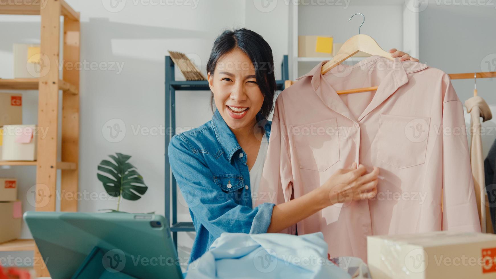 ung asiatisk dammodedesigner som använder mobiltelefon tar emot inköpsorder och visar kläder som spelar in video live streaming online i butik. småföretagare, online marknadsföringskoncept. foto
