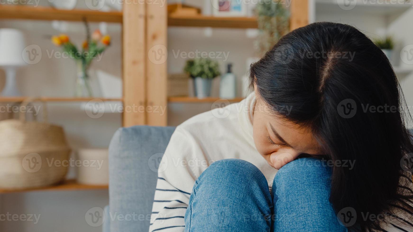 tankeväckande asiatisk dam sitter och omfamnar knän i soffan i vardagsrummet hemma och tittar ut utanför med ensam känsla, ledsen deprimerad tonåring tillbringar tid ensam stannar hemma, social distans, coronavirus -karantän. foto