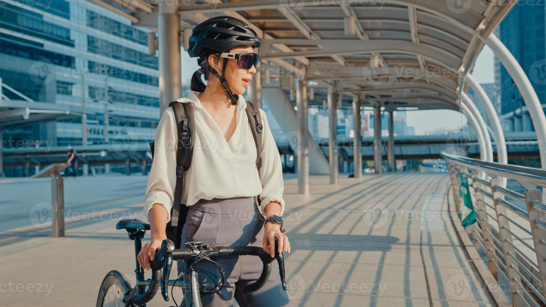 asiatisk dam affärskvinna bära solglasögon gå till jobbet på kontorspromenad och le titta runt håll cykelställ runt byggnaden på en stadsgata. cykelpendling, pendling på cykel, affärspendelkoncept. foto