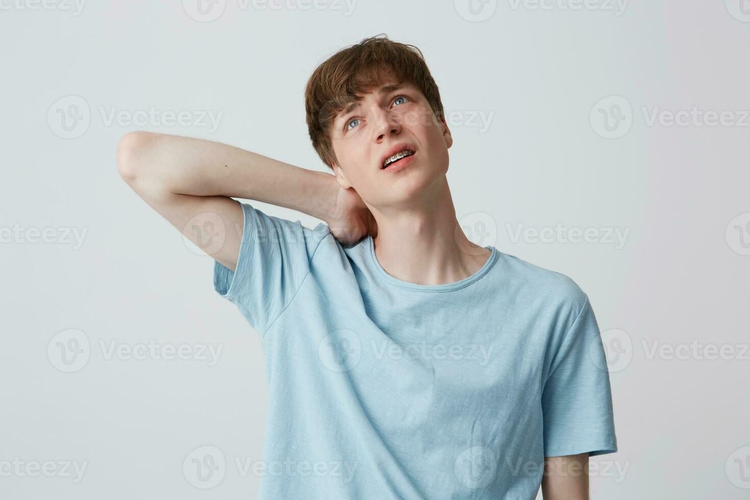 porträtt av ledsen desperat ung man med tandställning på tänder bär blå t skjorta känner trött och lidande från nacke smärta isolerat över vit bakgrund foto