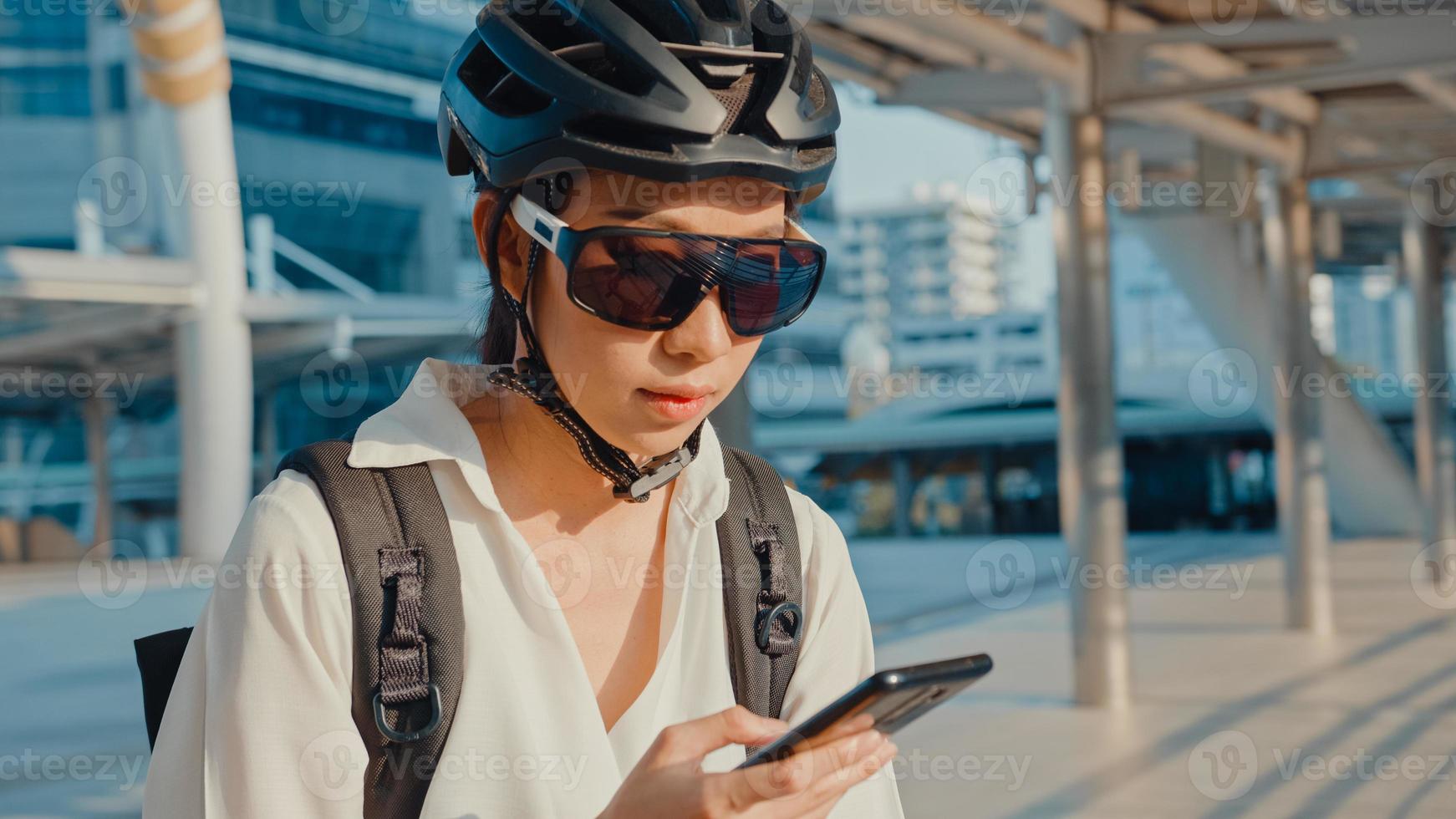 glad asiatisk affärskvinna med ryggsäck använder smart telefon i stadens stativ på gatan med cykel går till jobbet på kontoret. sportflicka använder sin telefon för att jobba. pendla till jobbet, affärspendlare i staden. foto