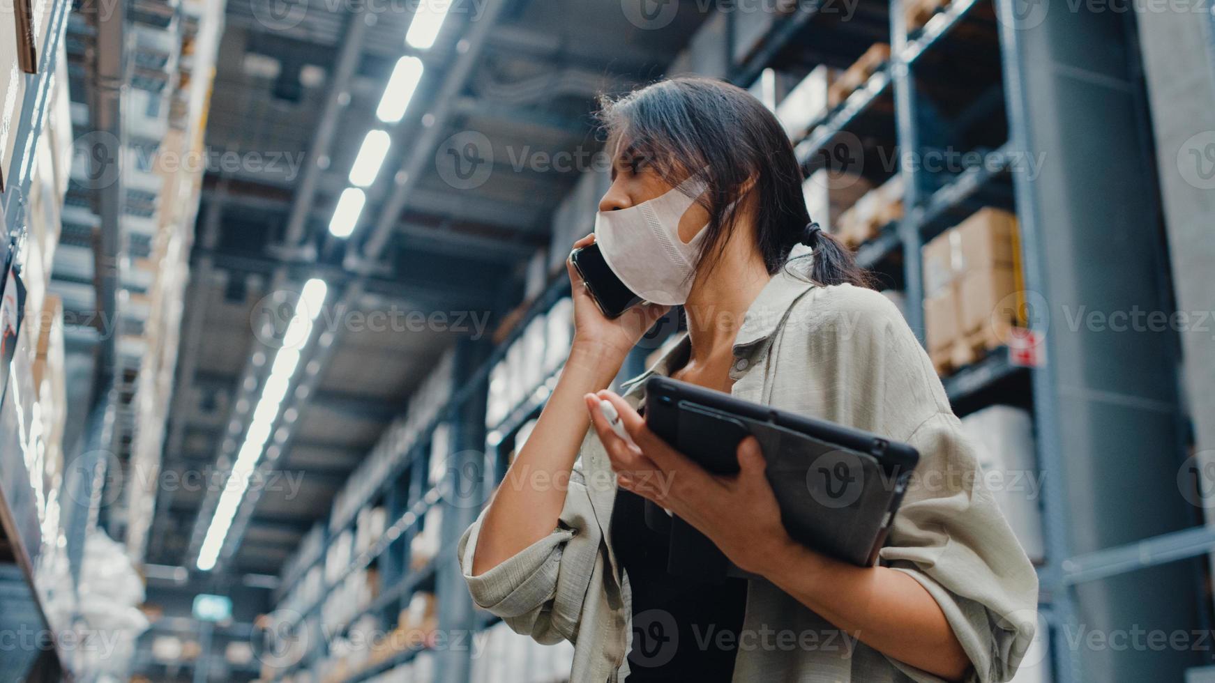 ung asiatisk affärskvinna online säljare bär ansiktsmask pratar bekräftar beställning från kund på telefon håll tablettställ i detaljhandeln köpcentrum. distribution, logistik, paket redo för leverans. foto