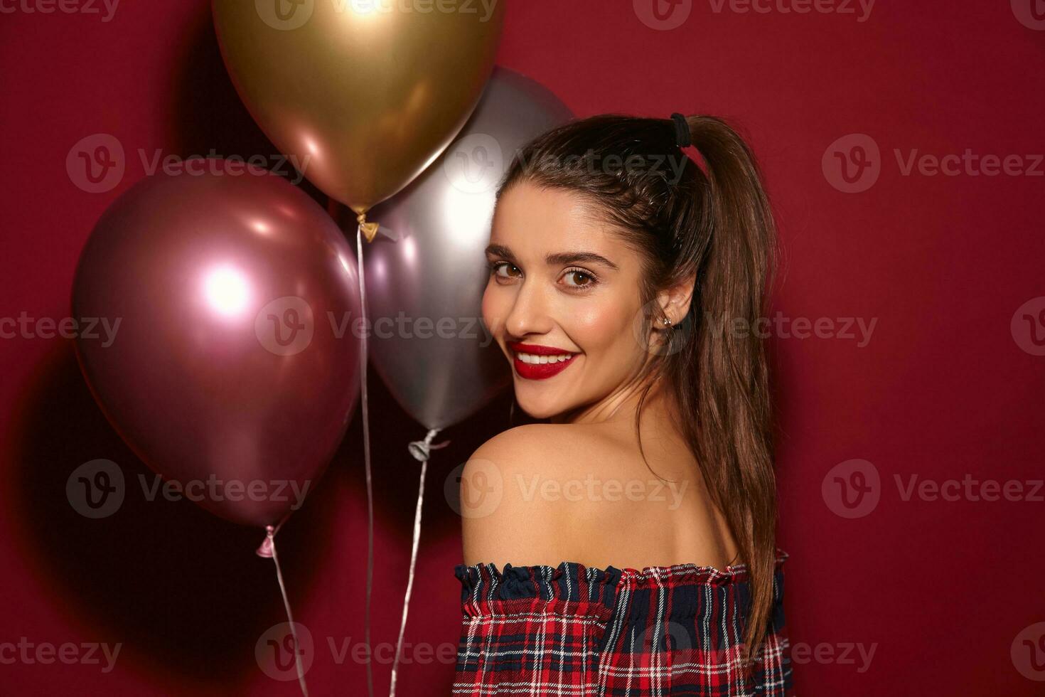 charmig ung positiv lång håriga brunett kvinna med tillfällig frisyr ser glatt över henne axel och leende brett, stående över vinröd bakgrund med luft ballonger foto