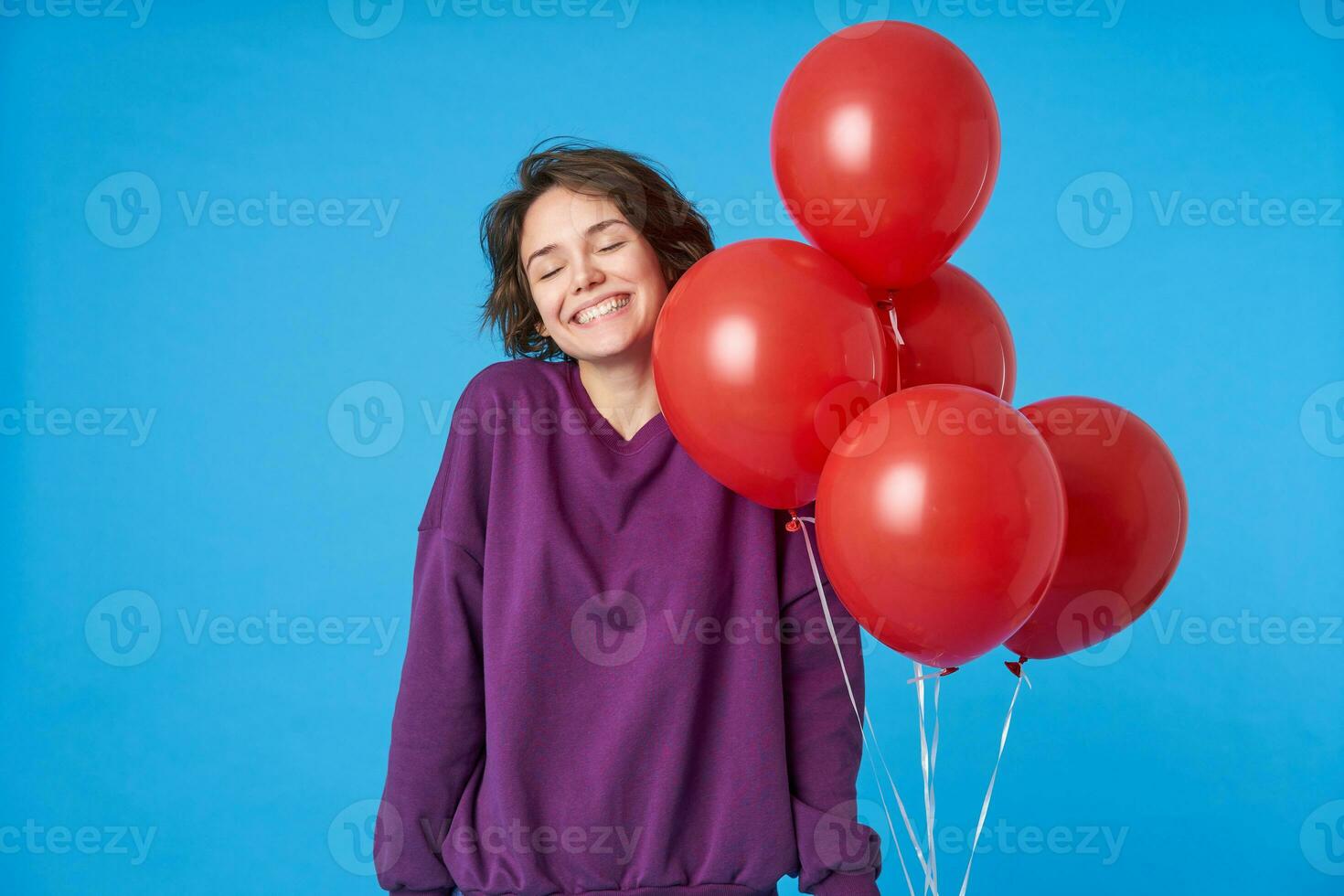 Bra ser ung glad mörk håriga kvinna med kort frisyr förvaring henne ögon stängd och leende allmänt, varelse överraskad med helium ballons förbi henne vänner, isolerat över blå bakgrund foto