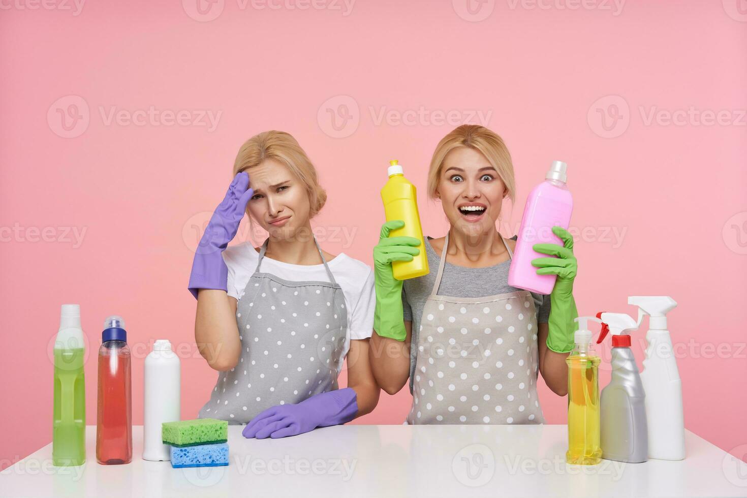 olycklig ung vithuvud rengöring lady ser tyvärr på kamera medan henne upphetsad blond flickvän höjning glatt henne händer med tvättmedel, isolerat över rosa bakgrund foto