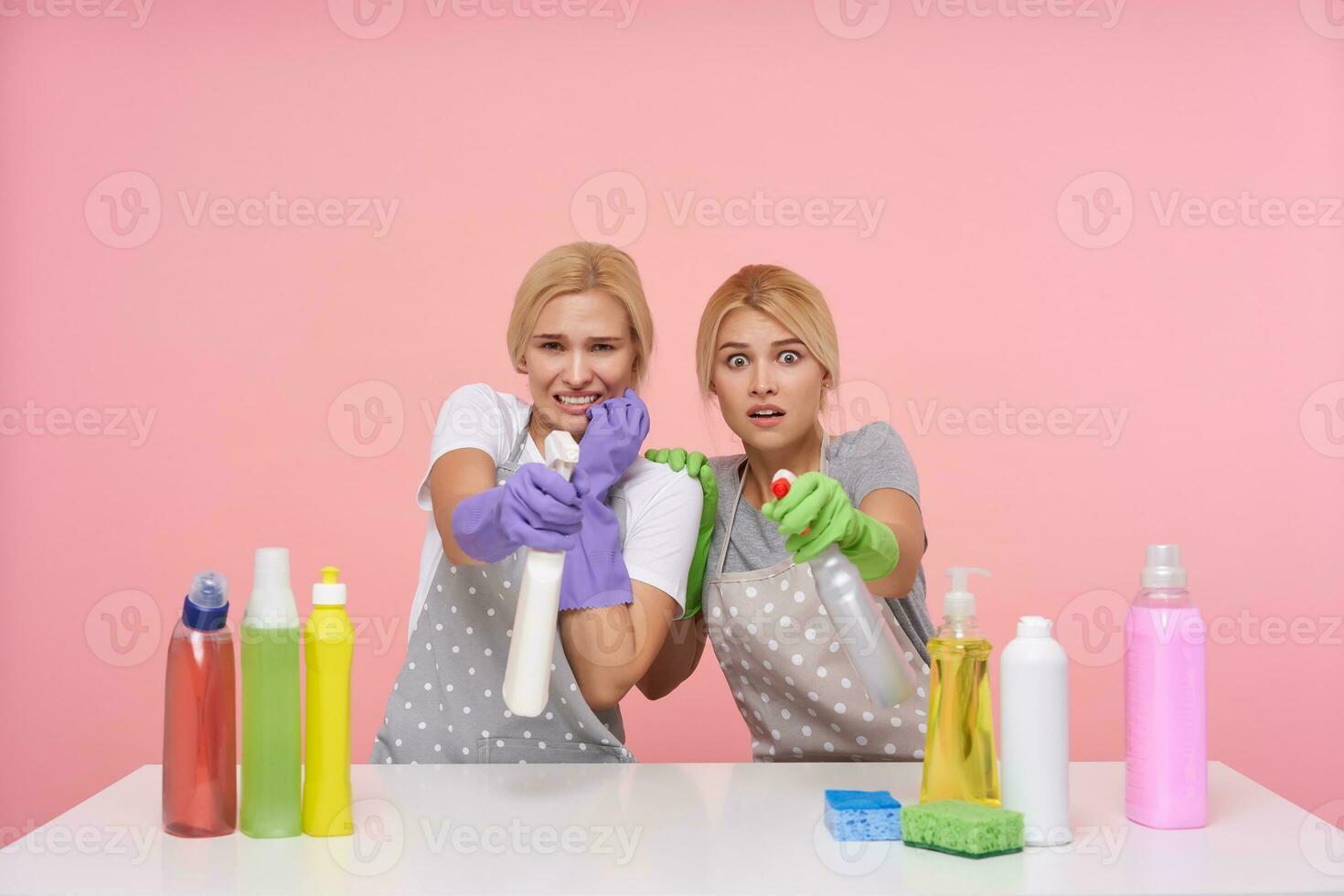 rädd ung vithuvud rengöring damer försvara sig själva med spray flaskor av tvättmedel, seende tjuvar medan framställning rengöring, Framställ över rosa bakgrund foto