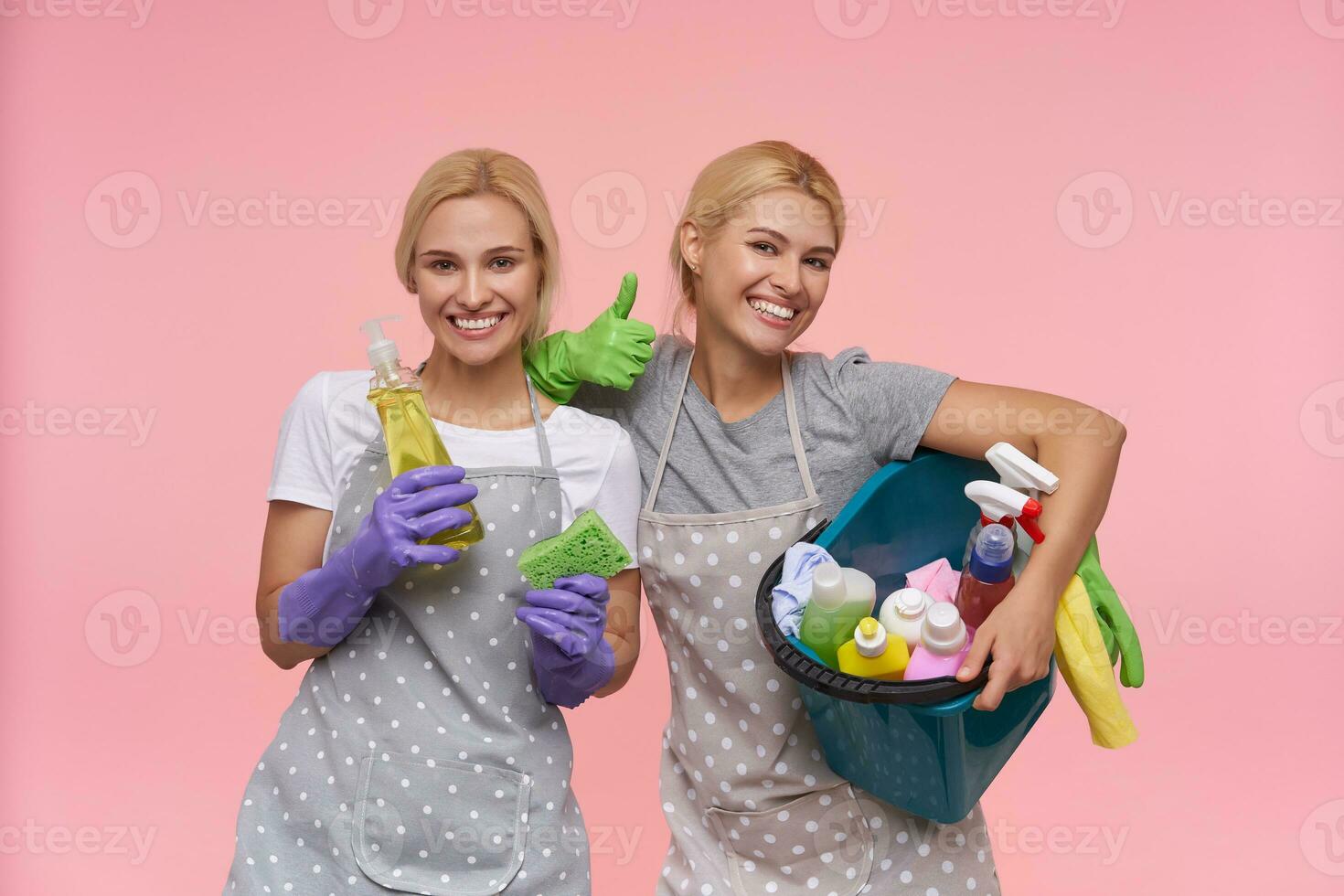 Lycklig ung Söt blond systrar med naturlig smink fröjd medan efterbehandling vår rengöring deras hus och Framställ över rosa bakgrund med hushåll kemikalier foto