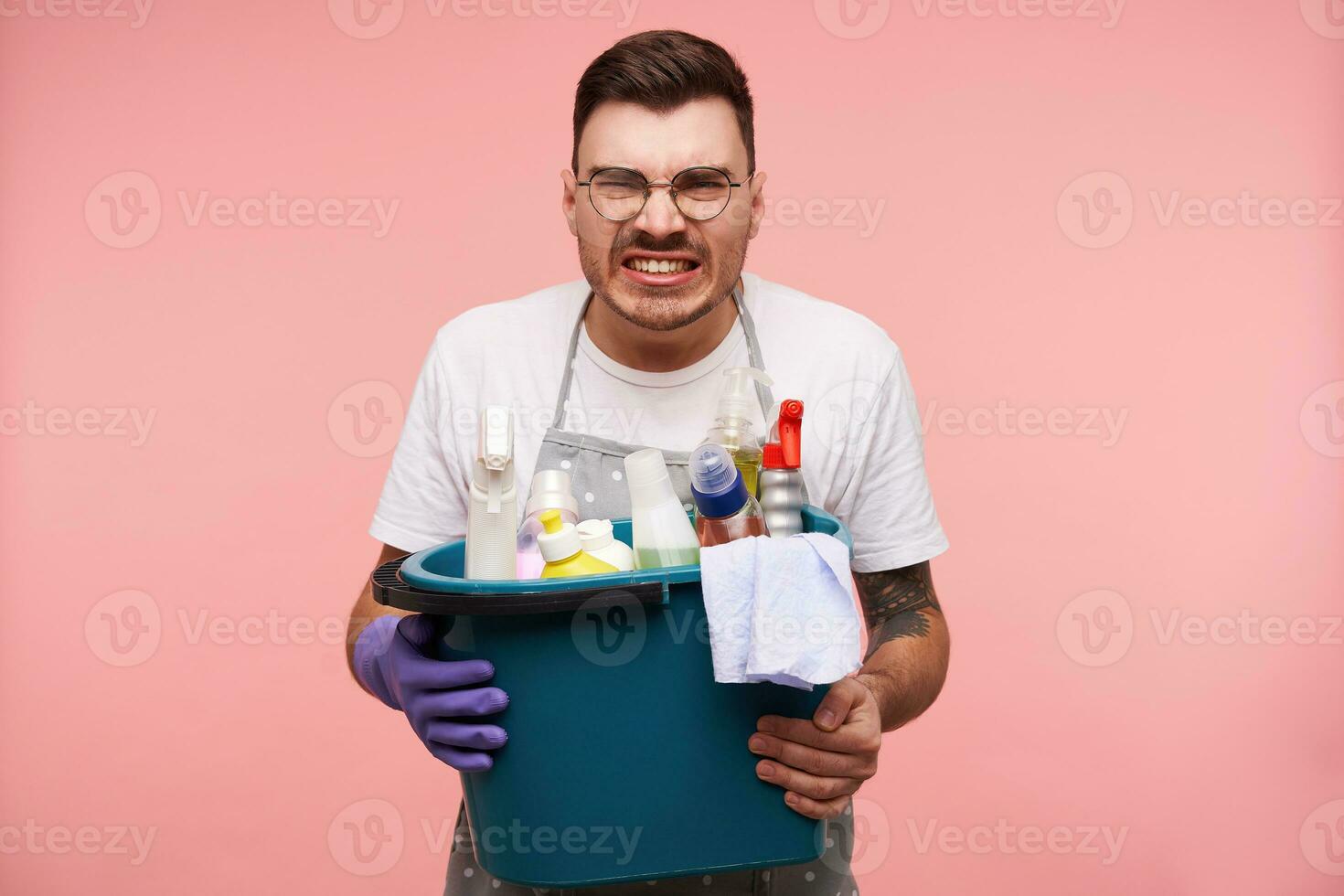 inomhus- skott av Söt ung brunett man med kort frisyr som visar hans tänder medan rynkar ansikte, bär arbetssätt kläder medan stående över rosa bakgrund foto