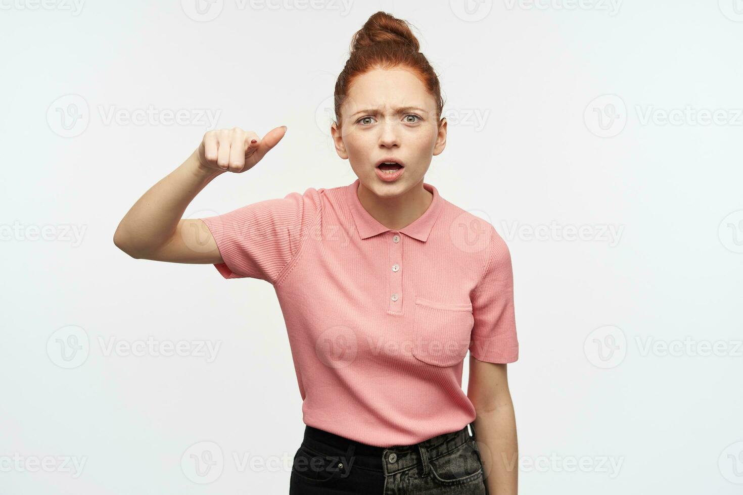 argumenterar kvinna, olycklig rödhårig flicka med hår bulle. bär rosa t-shirt och svart jeans. pekande med finger på du, skylla. tittar på på de kamera, isolerat över vit bakgrund. foto