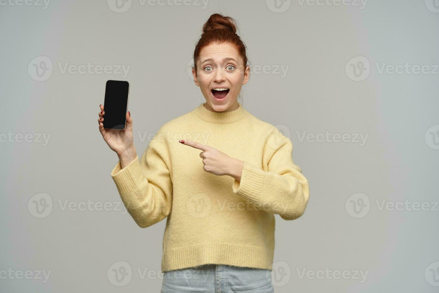 trevlig ser kvinna, glad rödhårig flicka med hår bulle. bär gul Tröja och innehav en smartphone. pekande på en skärm, på kopia klistra. tittar på på de kamera, isolerat över grå bakgrund foto