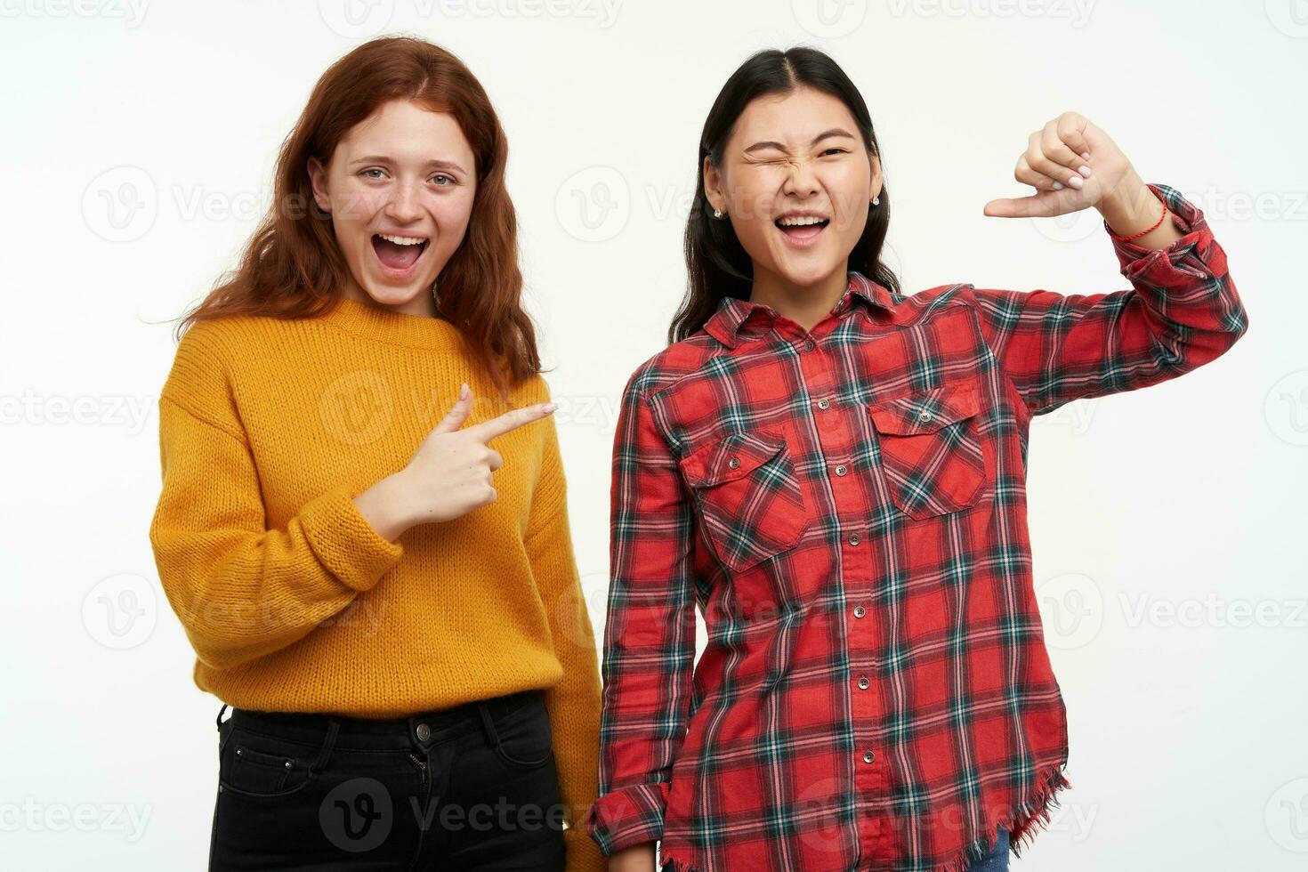 porträtt av glad asiatisk och caucasian vänner. bär tillfällig utrusta. Lycklig flicka pekande på henne vän vem blinkar och pekande på henne själv. tittar på på de kamera, isolerat över vit bakgrund foto
