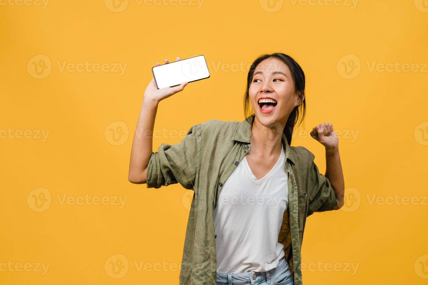 ung asiatisk dam visar tom smartphone -skärm med positivt uttryck, ler brett, klädd i vardagskläder som känner lycka på gul bakgrund. mobiltelefon med vit skärm i kvinnlig hand. foto