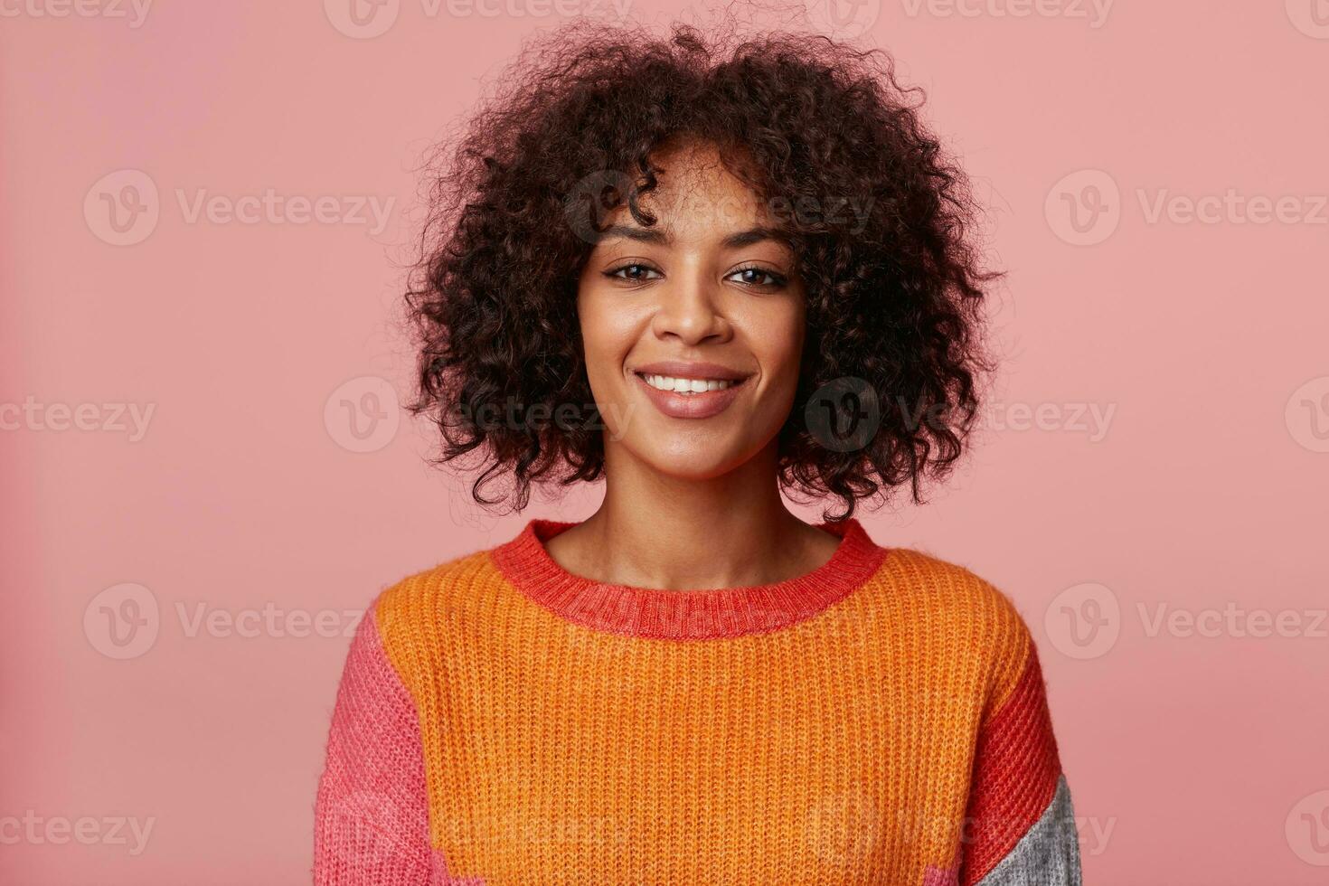 stänga upp porträtt av positiv optimistisk charmig afrikansk amerikan flicka med afro frisyr utseende med nöje, med vänlig leende, bär färgrik långärmad, isolerat på rosa bakgrund foto