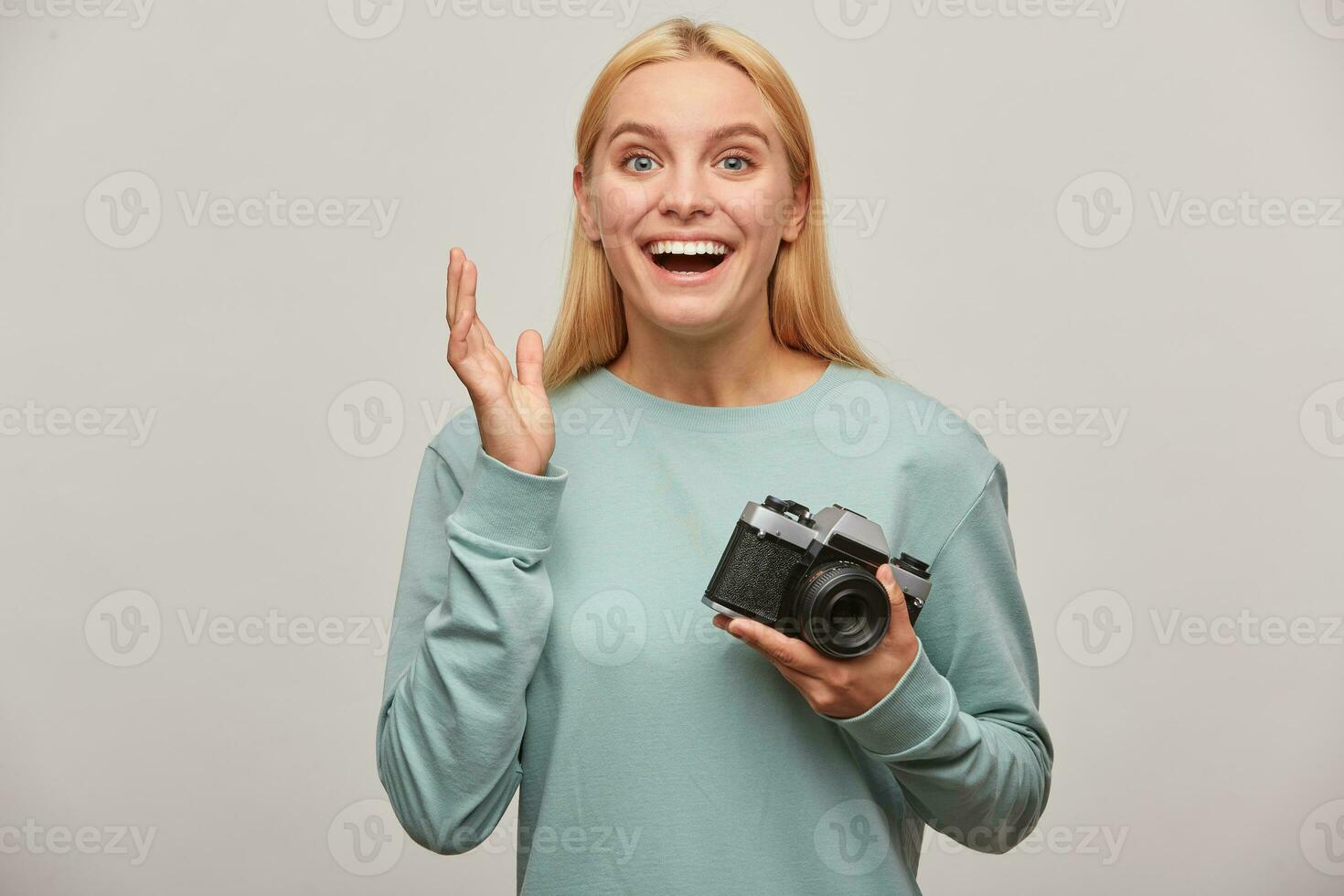 blond flicka fotograf, tar en Foto session, gjorde inte förvänta till skaffa sig så många bra skott, inspirerad förbi de retro årgång Foto kamera i hand, klädd i blå tröja, på grå bakgrund