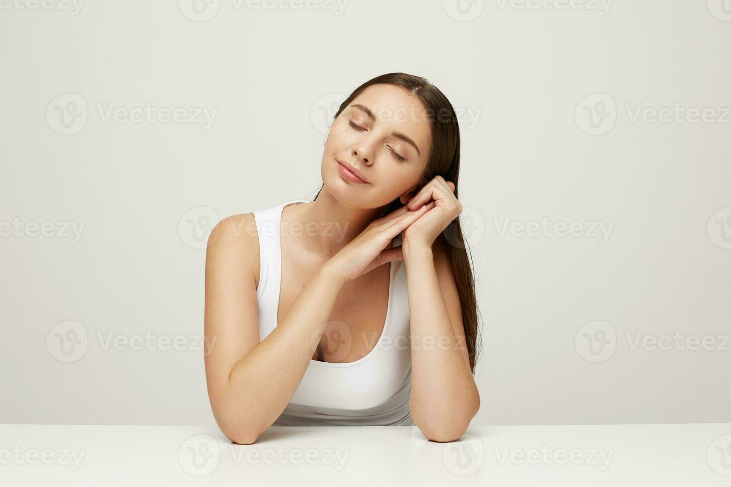 kvinna med perfekt friska färsk hud sitter på de tabell, händer sätta tillsammans nära ansikte och huvud lögner på dem, nöjd med de effekt av mask, ögon stängd i nöje, över vit bakgrund foto