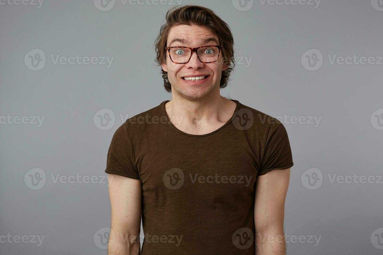 Foto av ung förvirrad man med glasögon bär i grundläggande t-shirt, står över grå bakgrund, Prova brett ler på de kamera.