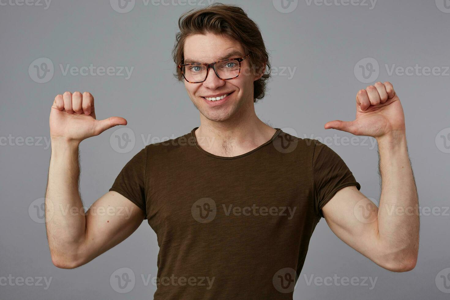 ung glad kille med glasögon bär i tom t-shirt stående över grå bakgrund och pekande på han själv, utseende Lycklig och brett ler och säger jag är Häftigt. foto