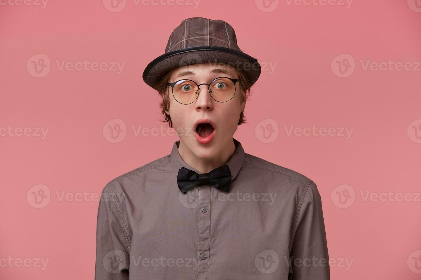 stänga upp av ung man med chockad, förvånad uttryck och tappade käke klädd i skjorta hatt och svart fluga bär glasögon känner överraskad isolerat på rosa bakgrund foto