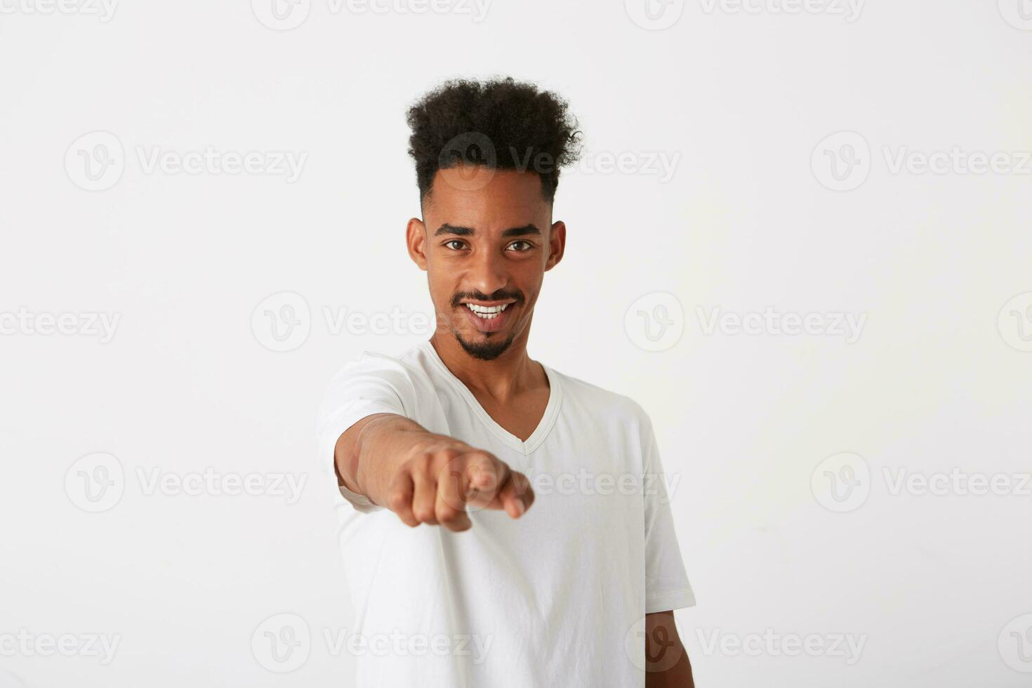 närbild av glad stilig afrikansk amerikan ung man med lockigt hår bär t skjorta utseende Lycklig och poäng på kamera isolerat över vit bakgrund foto