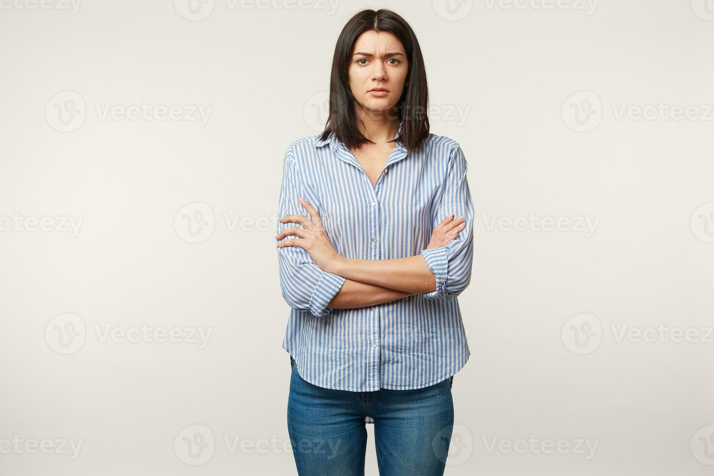 inomhus- Foto av brunett kvinna, utseende med misstro försiktig misstanke, spänd, lyssnar till någon med tvivel, stressig, stående med vapen vikta klädd i jeans och randig skjorta, över vit vägg