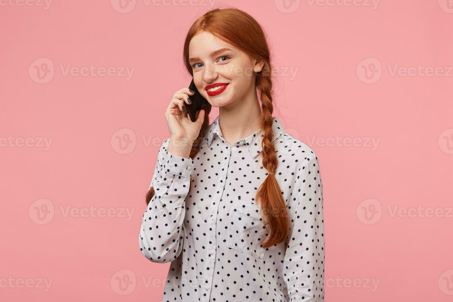 ung attraktiv rödhårig flicka med flätor klädd i en vit prickar skjorta innehav en telefon nära de öra med hand talande till någon ser på de kamera med le, isolerad på en rosa bakgrund foto