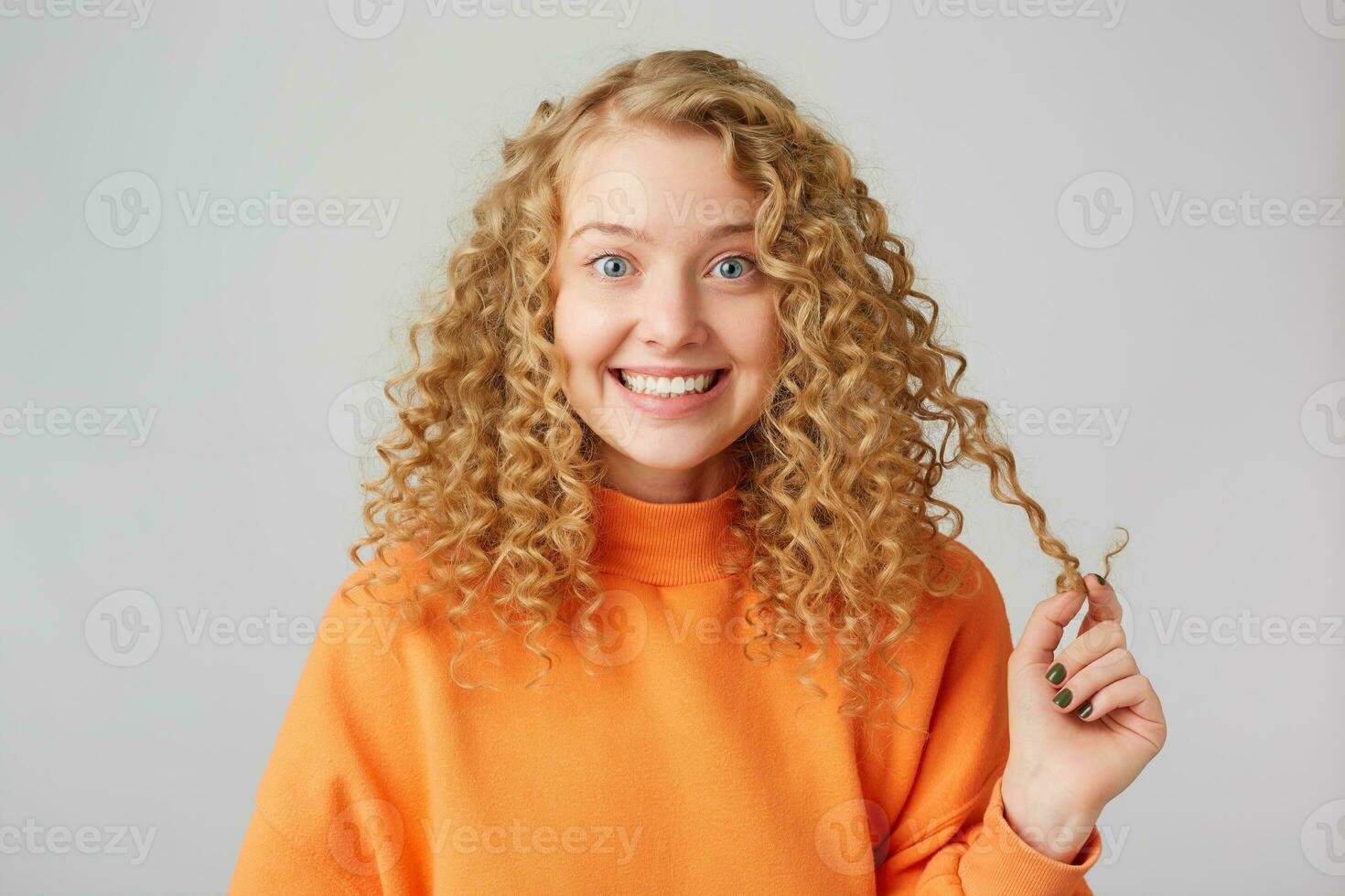 vänlig attraktiv glad ung blond kvinna ler vänliga hälsningar, nöjd, i en Bra humör, tittar på de kamera, håller en strå av hår klädd i ett orange Tröja, isolerat mot en vit bakgrund foto