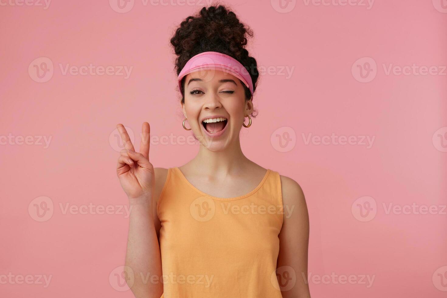 Tonårs flicka, flörtig ser kvinna med mörk lockigt hår bulle. bär rosa visir, örhängen och orange tank topp. har göra upp. visar fred sjunga. blinkar på de kamera isolerat över pastell rosa bakgrund foto