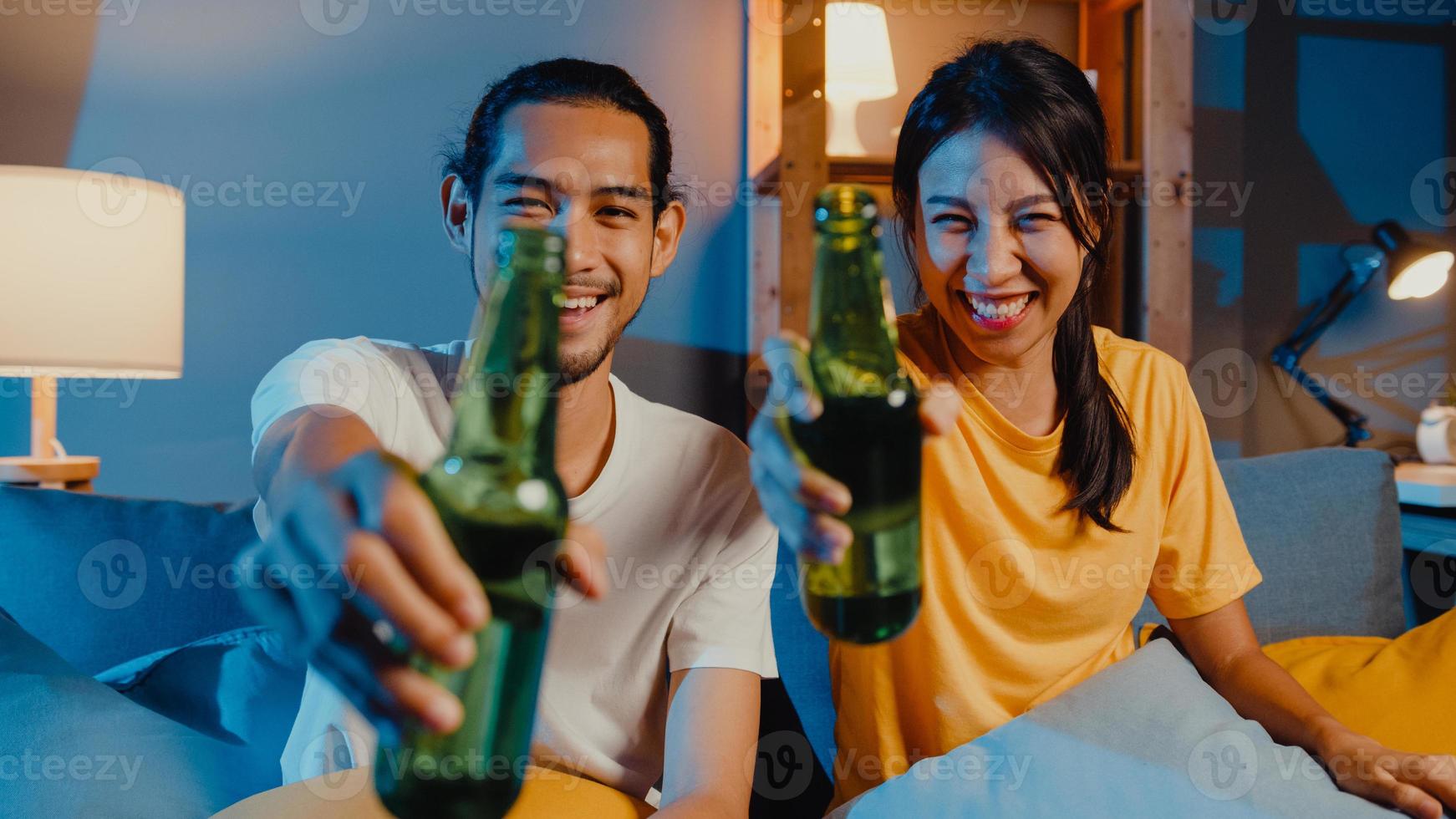 glada unga asiatiska par tittar på kameran njuta av nattfest händelse online sitta soffan videosamtal med vänner toast dricka öl via videosamtal online i vardagsrummet hemma, social distansering koncept. foto