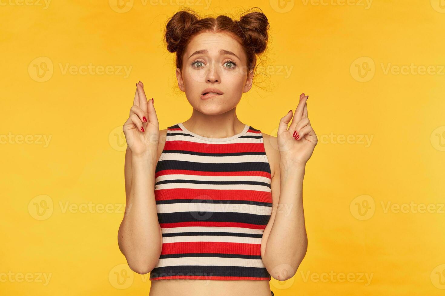 porträtt av attraktiv, röd hår flicka med två bullar. bär randig tank topp och korsa henne fingrar för en önskar, chockad, bita henne läpp. tittar på på de kamera isolerat över gul bakgrund foto