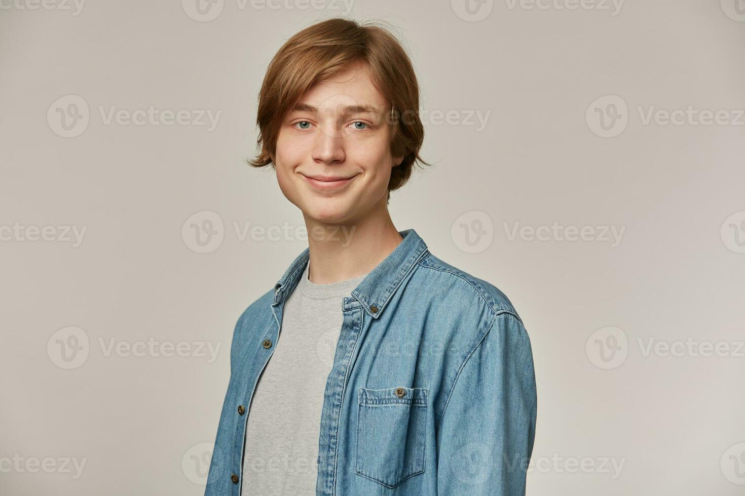 positiv Tonårs kille, Lycklig ser man med blond hår. bär blå denim skjorta. människor och känsla begrepp. tittar på och leende på de kamera isolerat över grå bakgrund foto