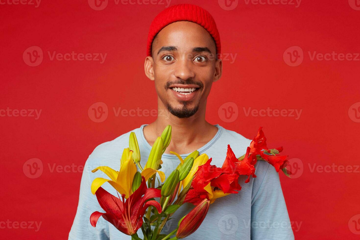 porträtt av ung glad attraktiv kille i röd hatt och blå t-shirt, innehar en bukett i hans händer, utseende på de kamera med Lycklig uttryck och leende, står över röd bakgrund. foto