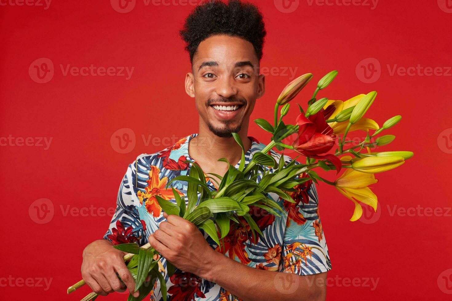 porträtt av ung leende afrikansk amerikan man, bär i hawaiian skjorta, utseende på de kamera med Lycklig uttryck, innehar gul och röd blommor bukett , står över röd bakgrund. foto