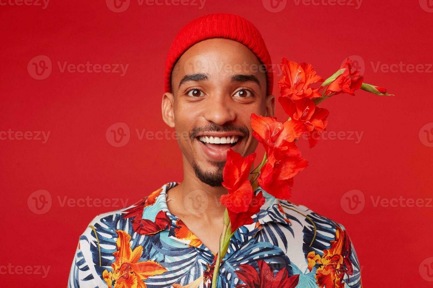 Foto av ung attraktiv Lycklig mörk flådd man, bär i hawaiian skjorta och röd hatt, utseende på de kamera med Lycklig uttryck, står över röd bakgrund.